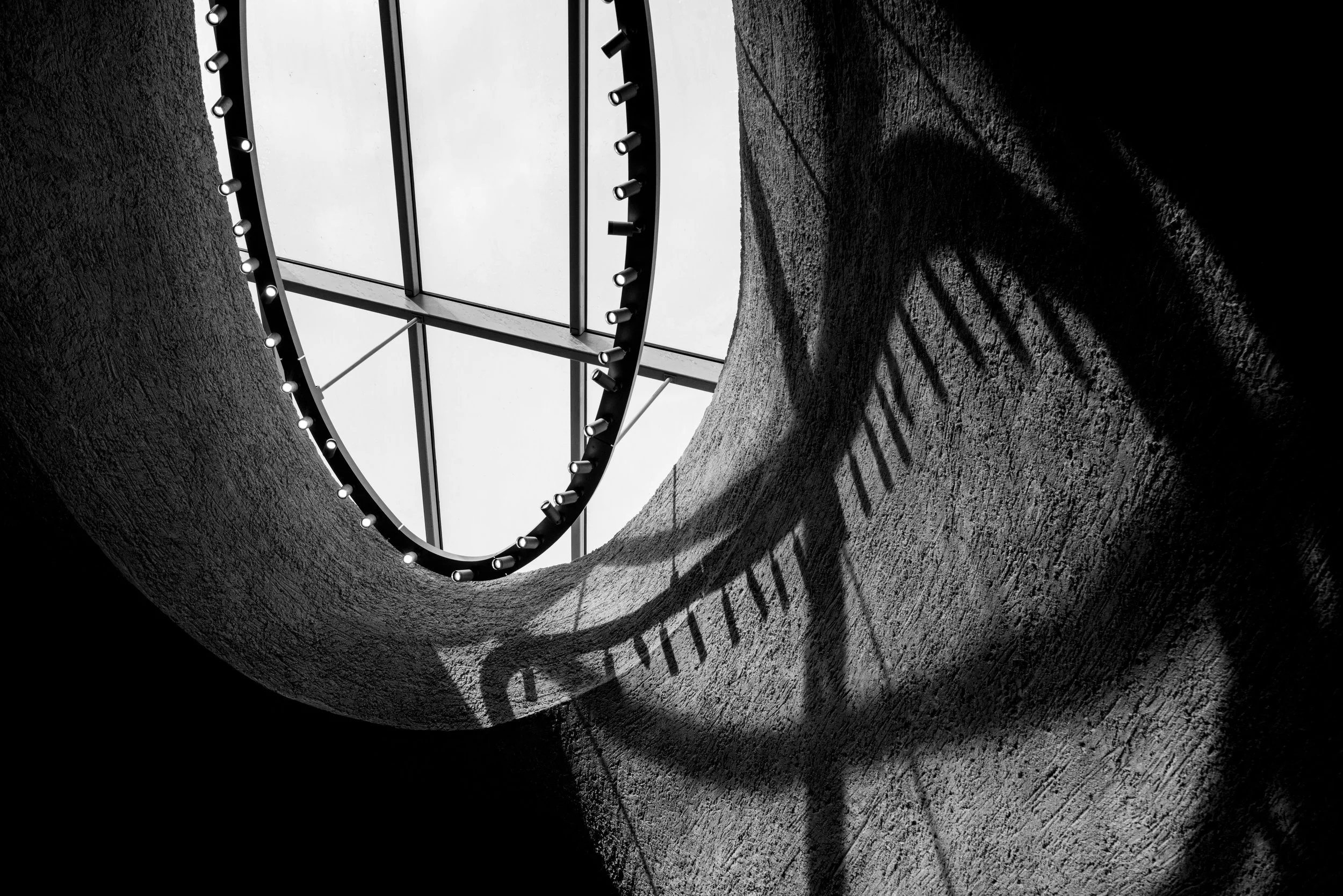 Black-and-white close-up of a curved architectural structure with rows of cylindrical lights beneath a glass canopy