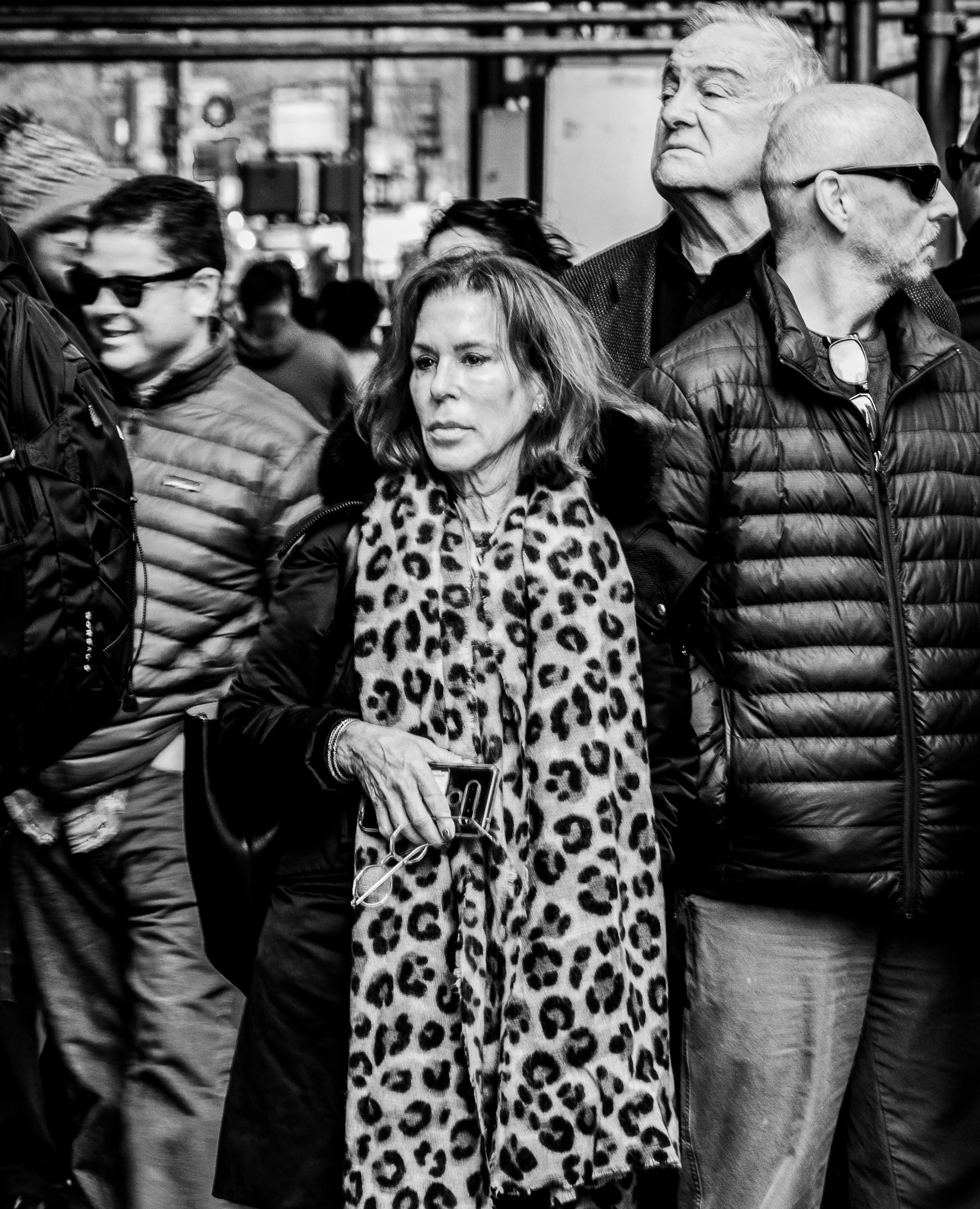 Black-and-white photo of a crowded group of people in winter clothing, including a woman in a leopard-print scarf and men wearing sunglasses.