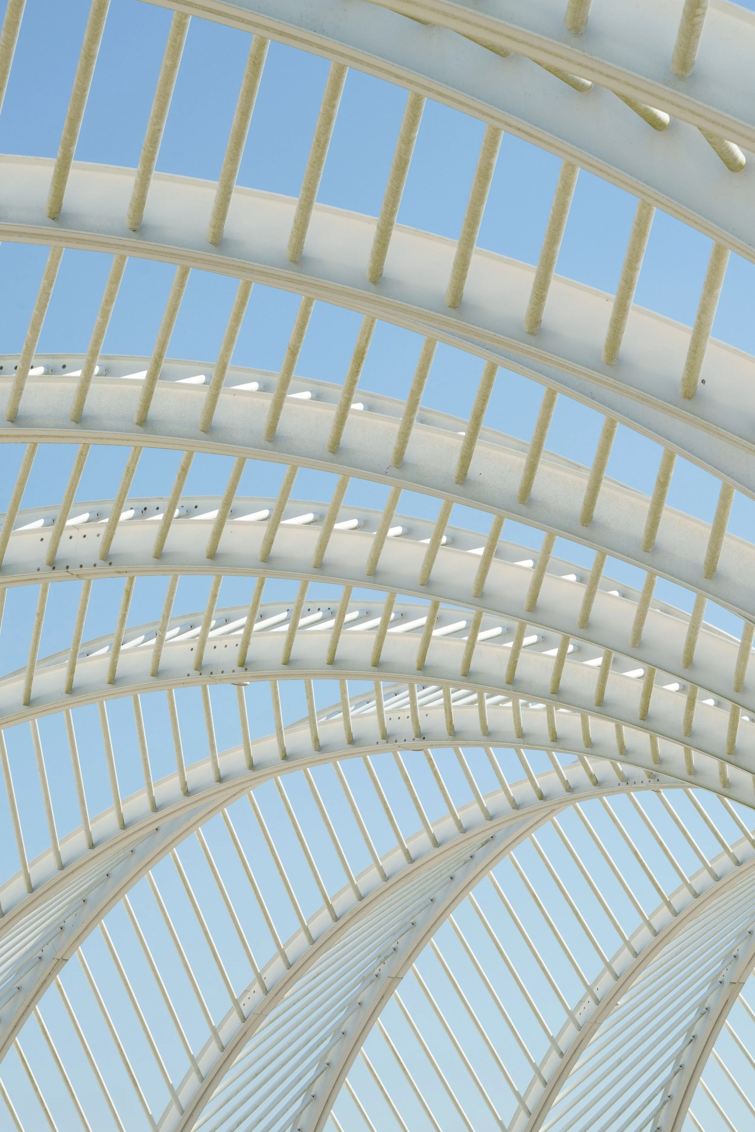 White curved architectural canopy with repeating beams and rods against a clear blue sky