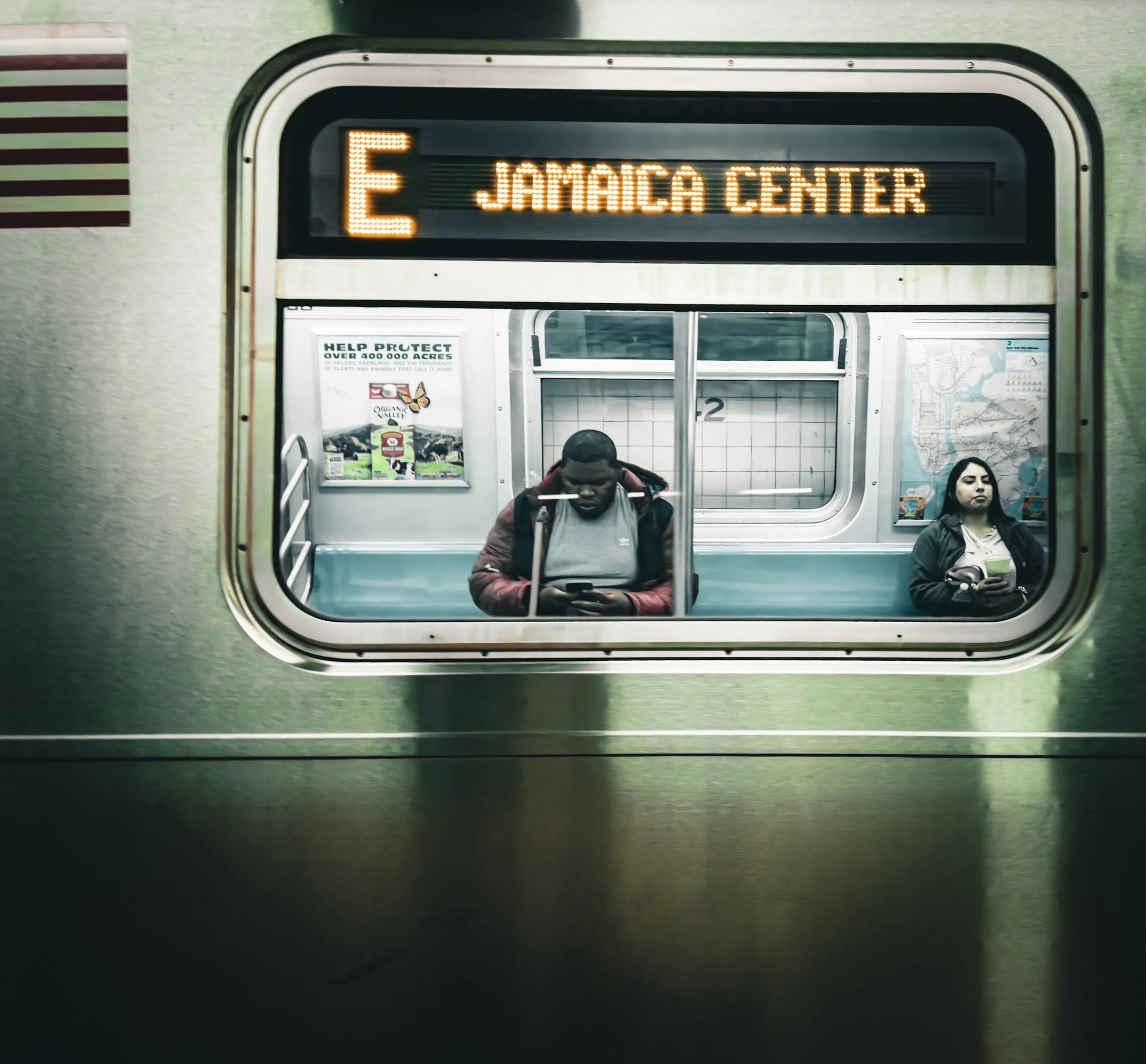 Subway train window showing two passengers and an illuminated Jamaica Center destination sign