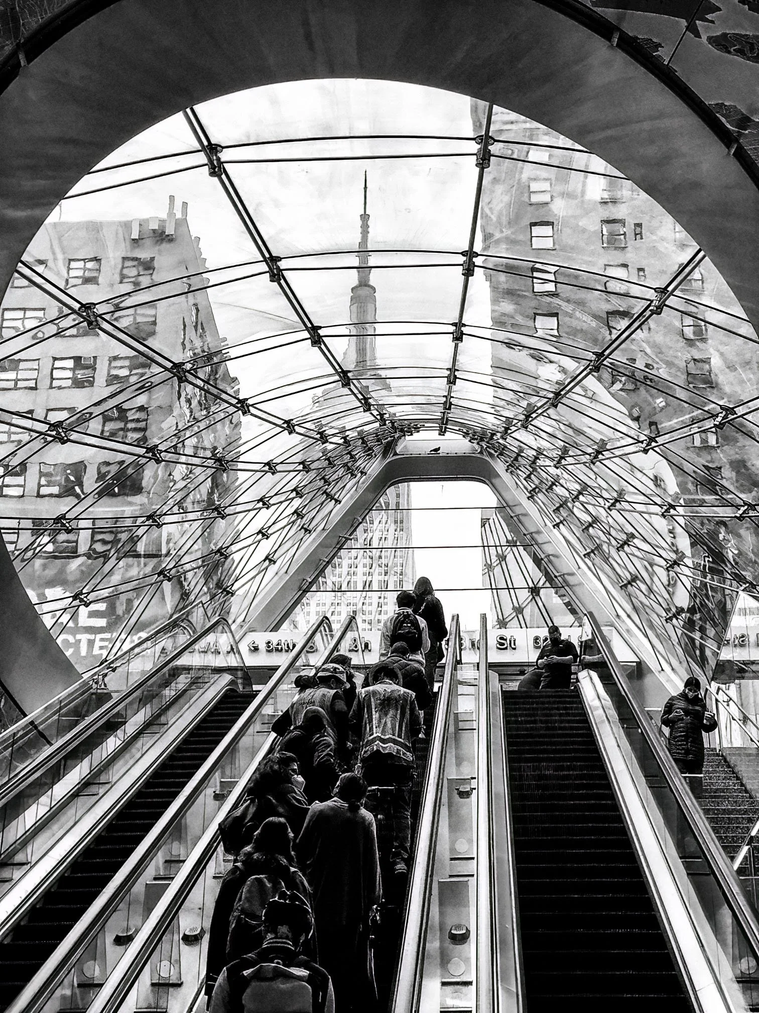 Black-and-white view of commuters riding escalators beneath a glass-and-steel canopy in a city subway entrance