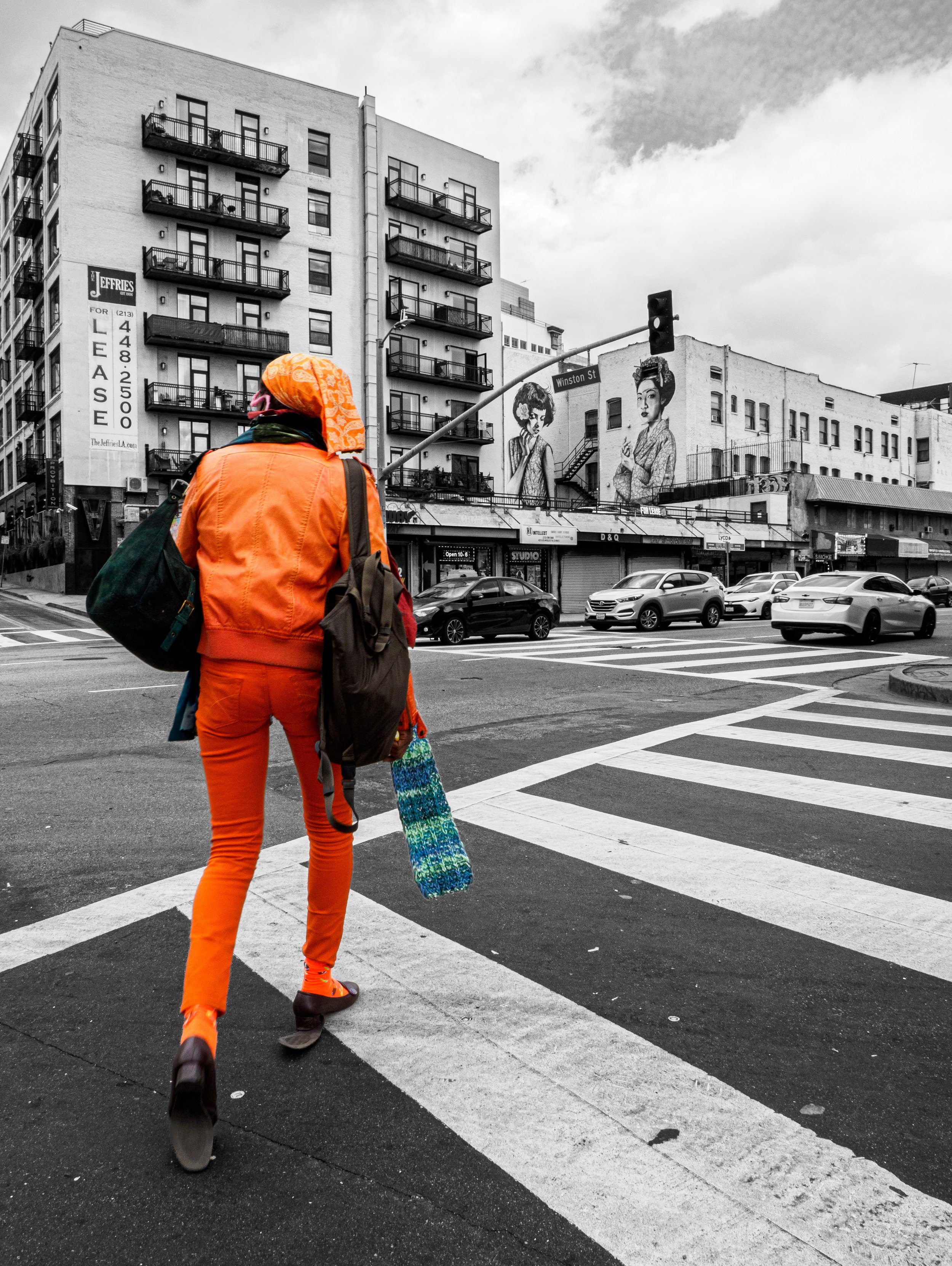 Person in bright orange clothing crossing a city intersection beneath the Winston Street sign, with urban buildings, murals, traffic lights, and cars in the background.