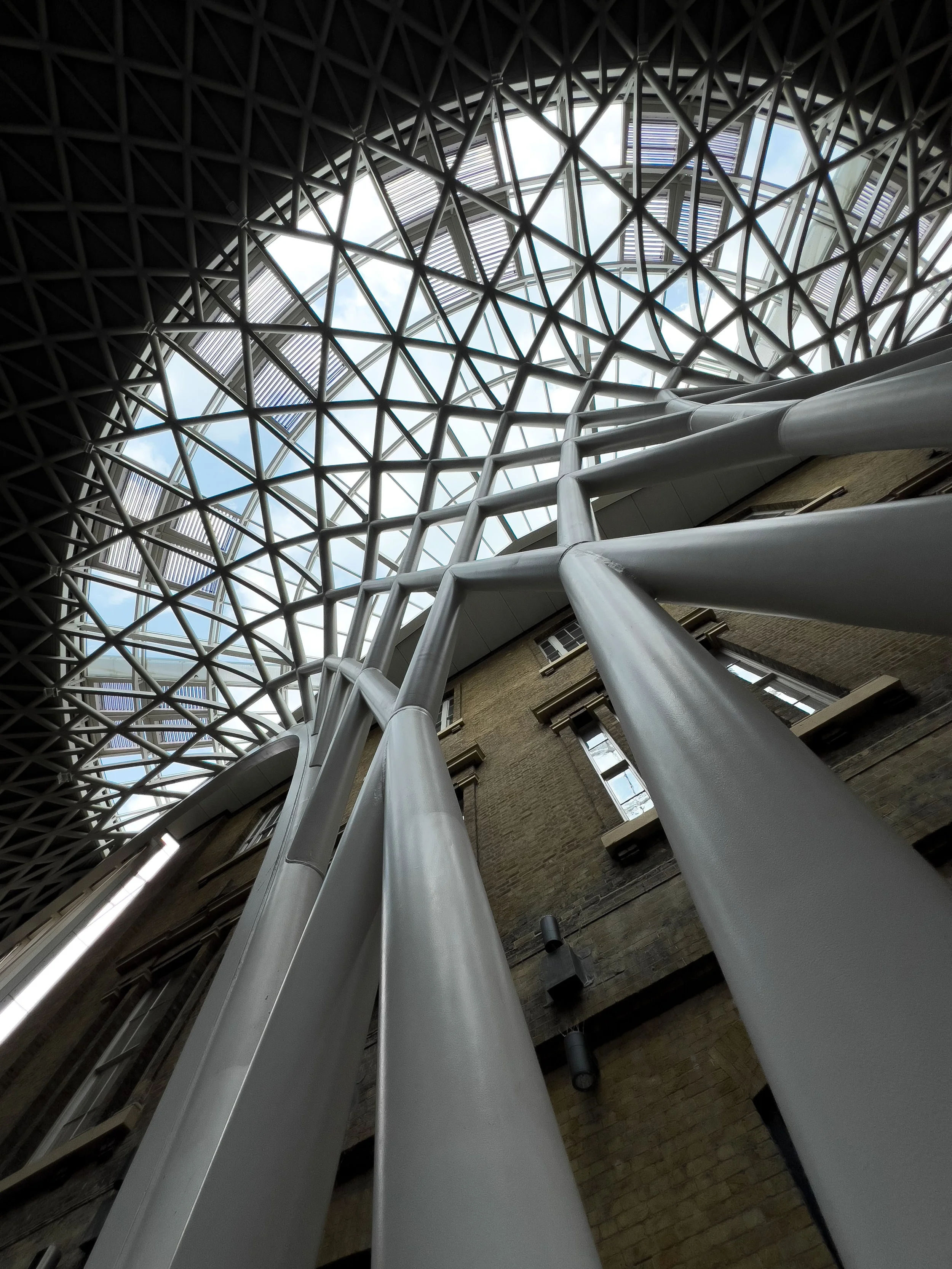Upward view of a modern atrium with white structural beams, a geometric glass skylight, and exposed brick walls.