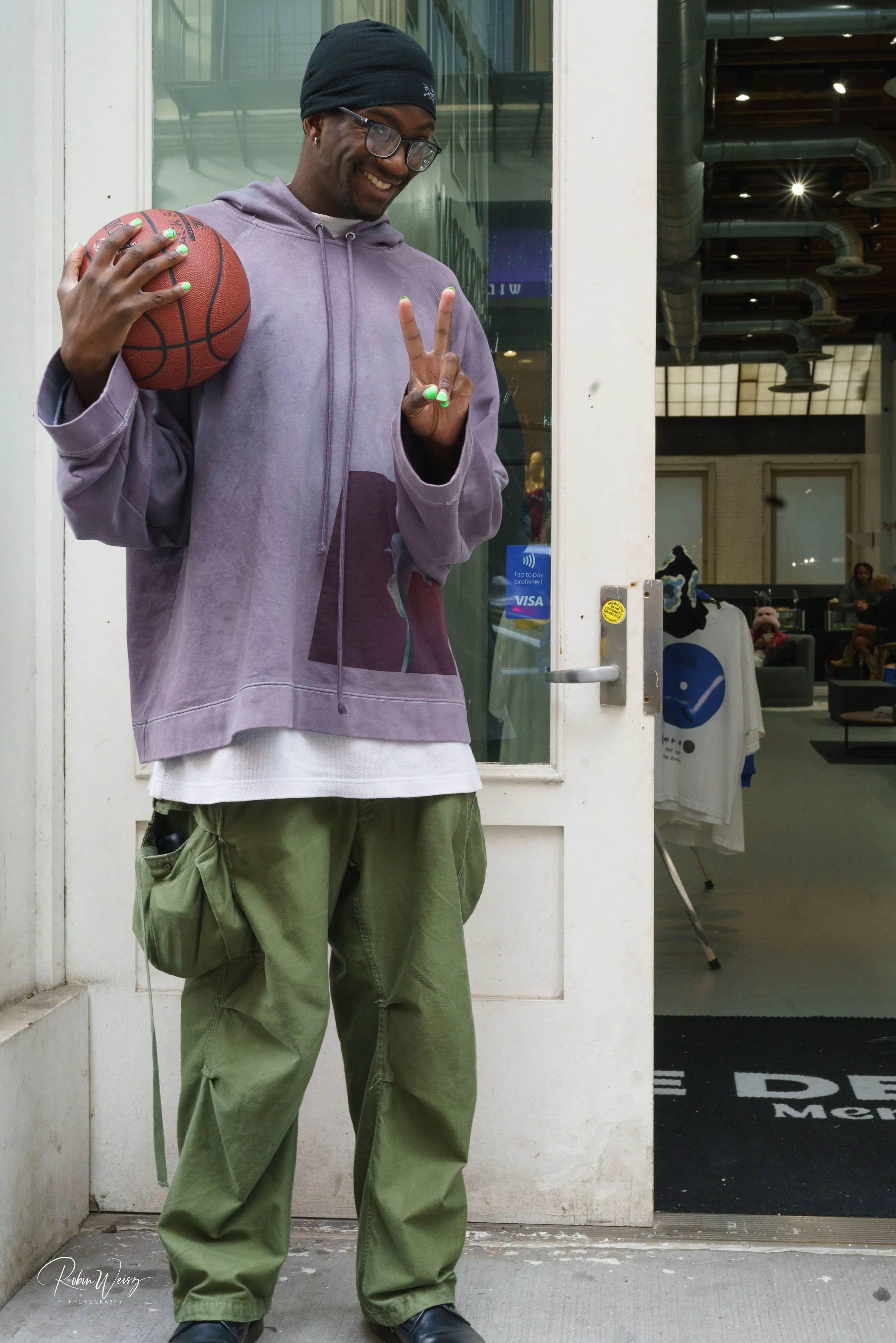 Smiling man in a purple hoodie and beanie holding a basketball and flashing a peace sign outside a storefront