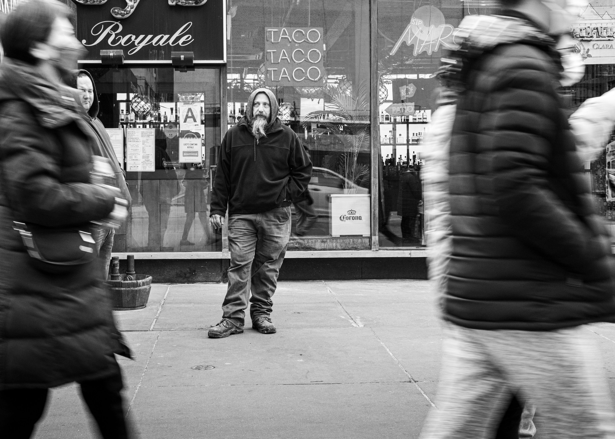 Black-and-white street photograph of a bearded man standing outside a taco restaurant as blurred pedestrians pass by.