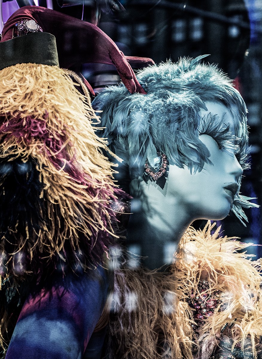 Fashion model wearing an avant-garde blue feathered headpiece, jeweled earring, and layered shaggy feather garments.