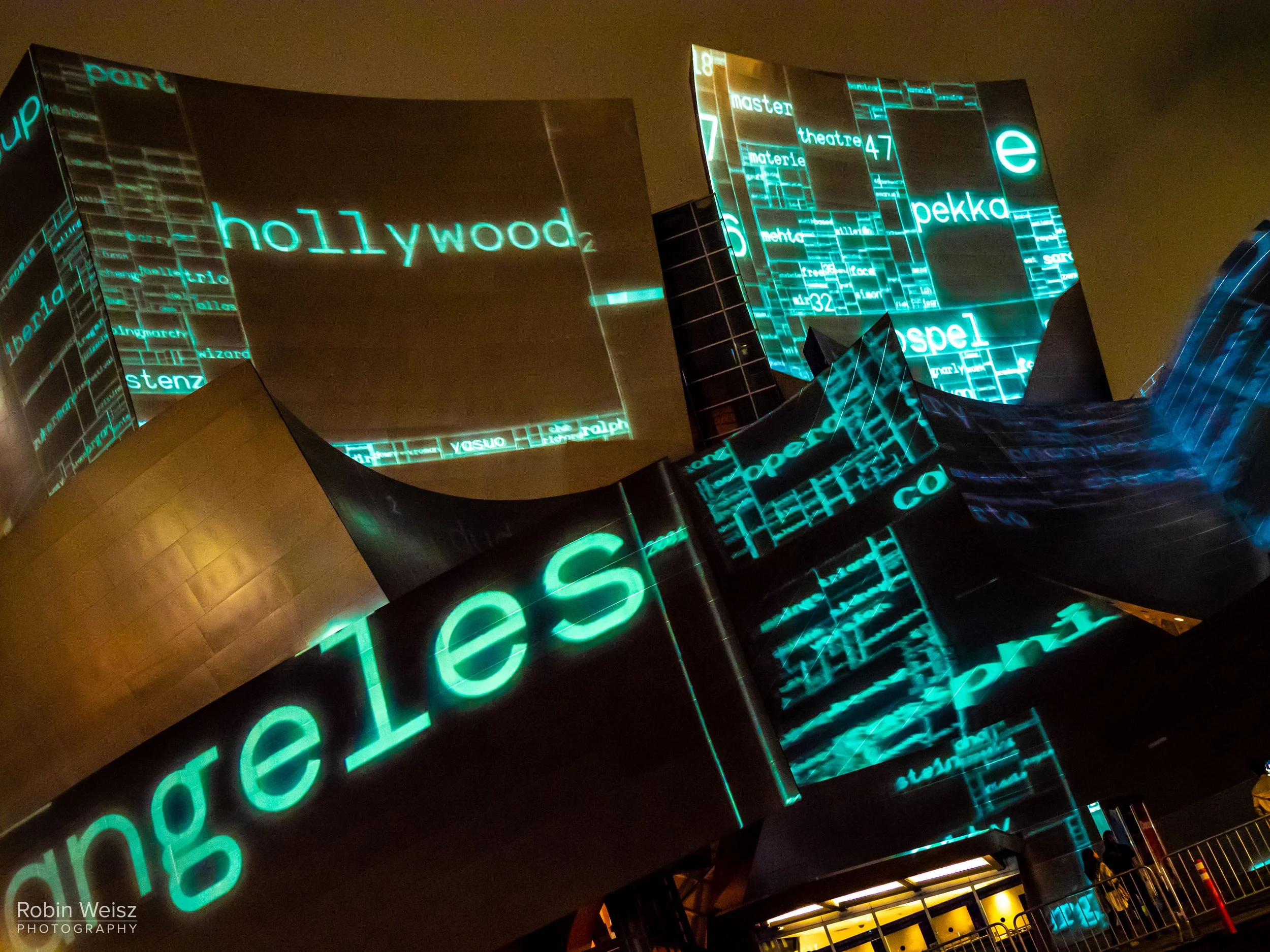 Nighttime abstract projection mapping displays glowing turquoise words and letters across the angular exterior of a modern building.
