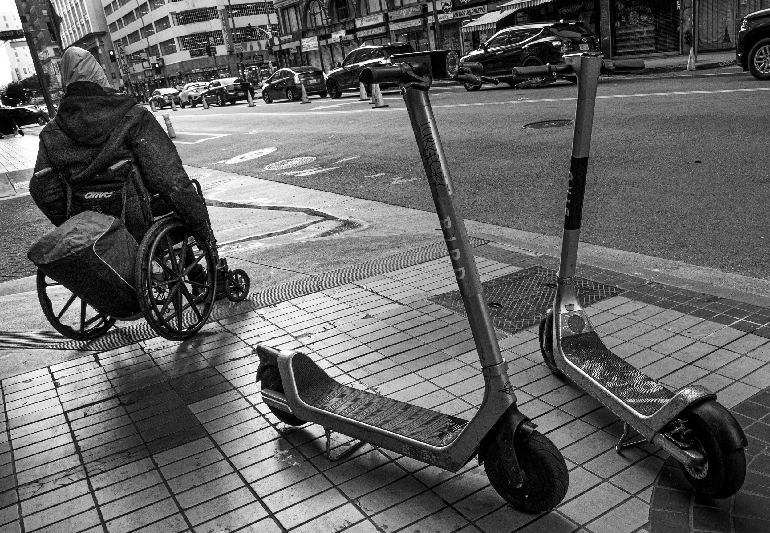Black-and-white city street scene with two electric scooters, a person in a wheelchair, parked cars, and storefronts.