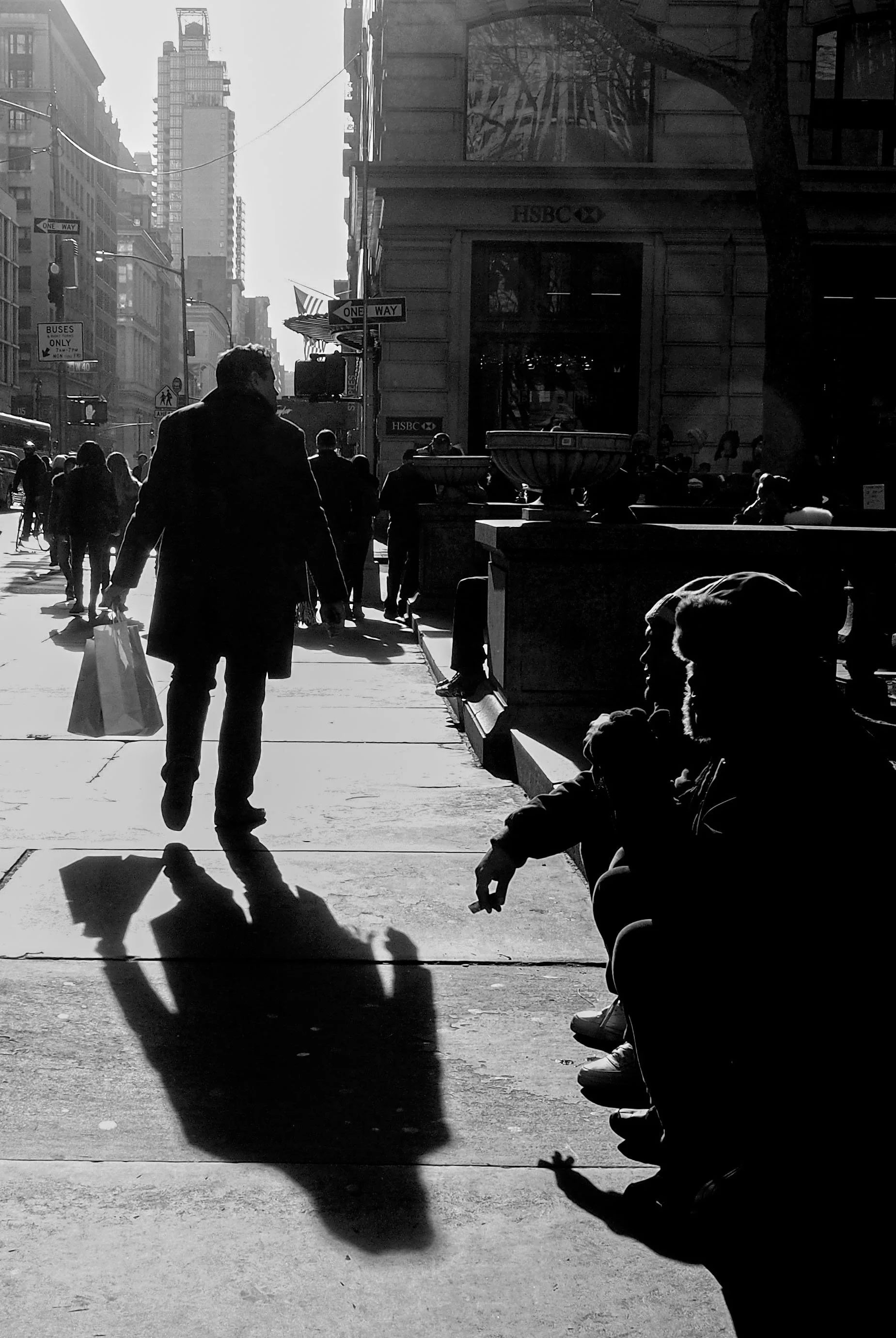 Black-and-white street scene with pedestrians, one-way signs, an HSBC bank, and long shadows on a sunny urban sidewalk.