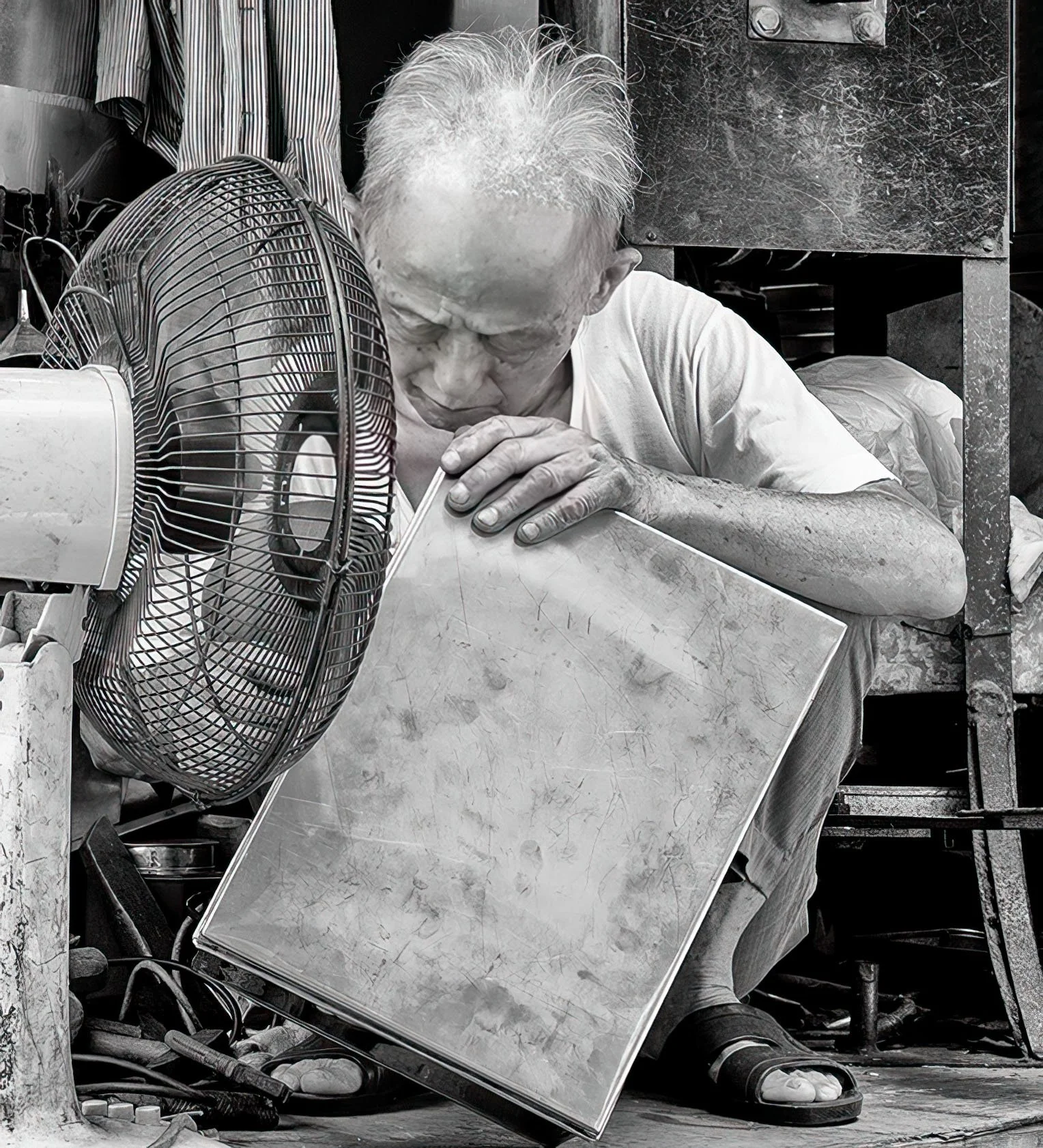 Black-and-white photo of an elderly man resting his head on a large sheet of metal beside a floor fan in a cluttered workshop.