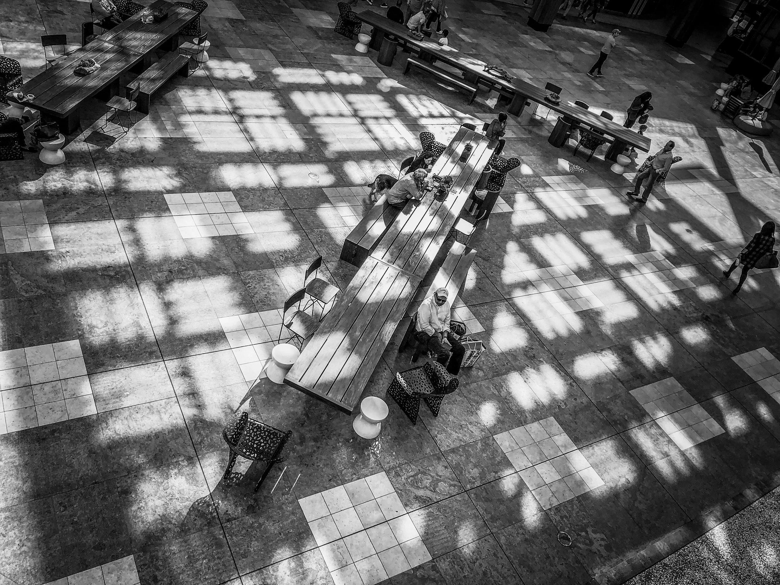 Black-and-white overhead view of people sitting at long communal tables in a spacious indoor food court.
