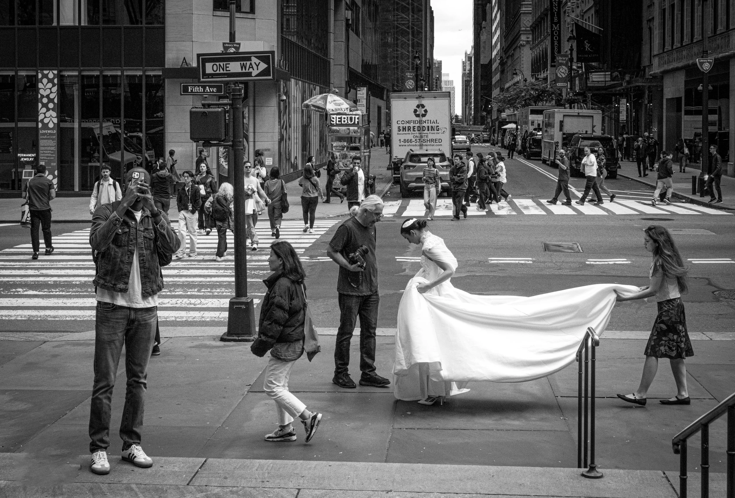 Black-and-white street scene at Fifth Avenue and Library Way in New York City, with pedestrians crossing and people carrying a large white fabric banner.