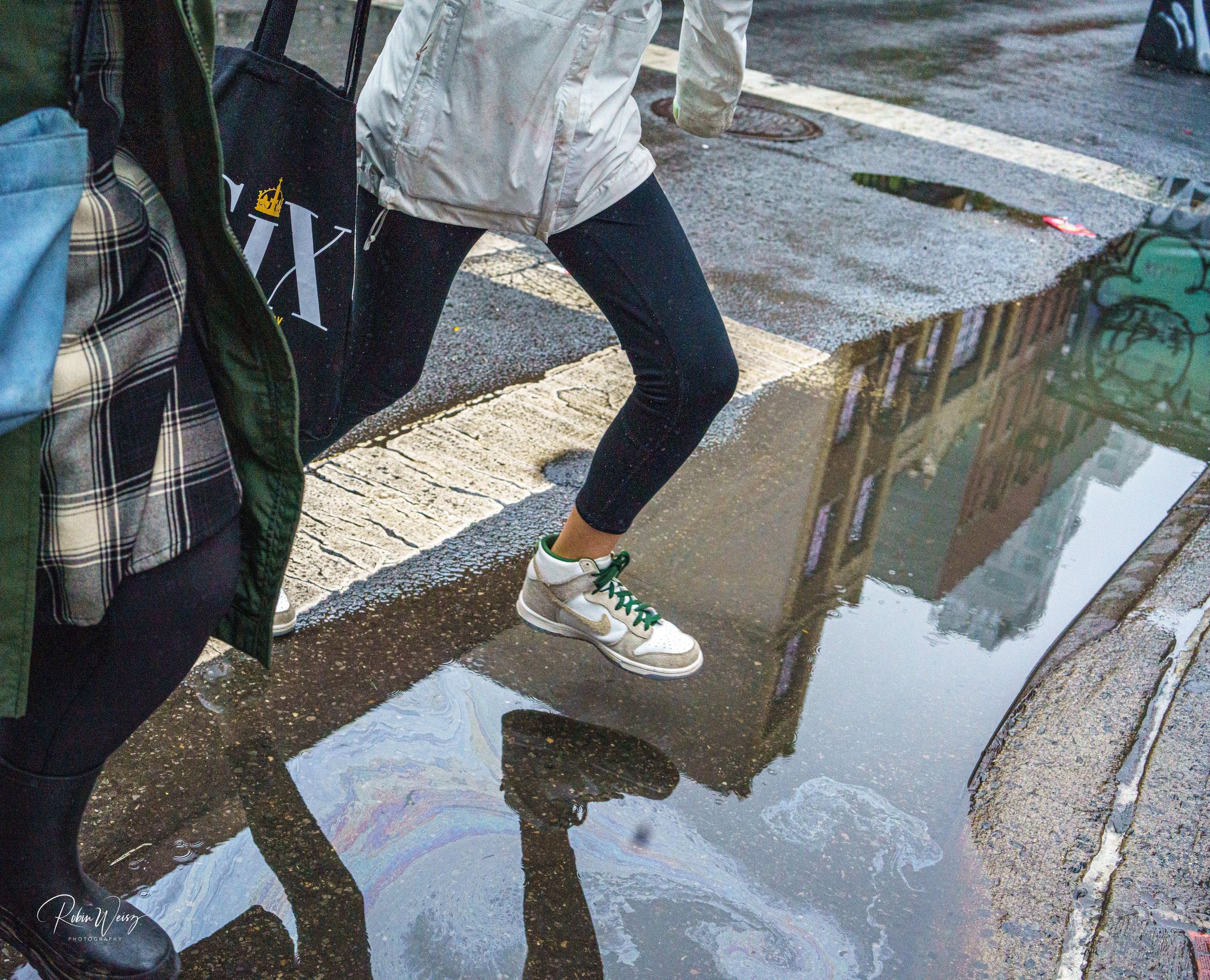 Person in a white jacket and black leggings stepping through a rain puddle beside a plaid suitcase and umbrella.