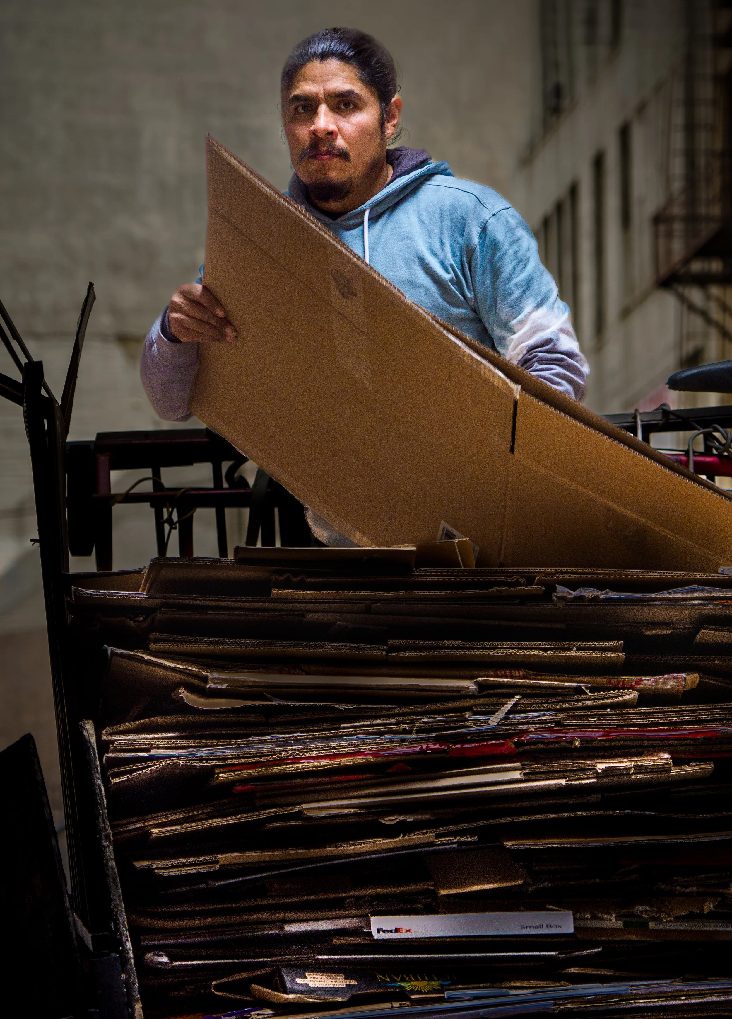 Man in a blue hoodie carrying a large cardboard box beside a stack of flattened cardboard.