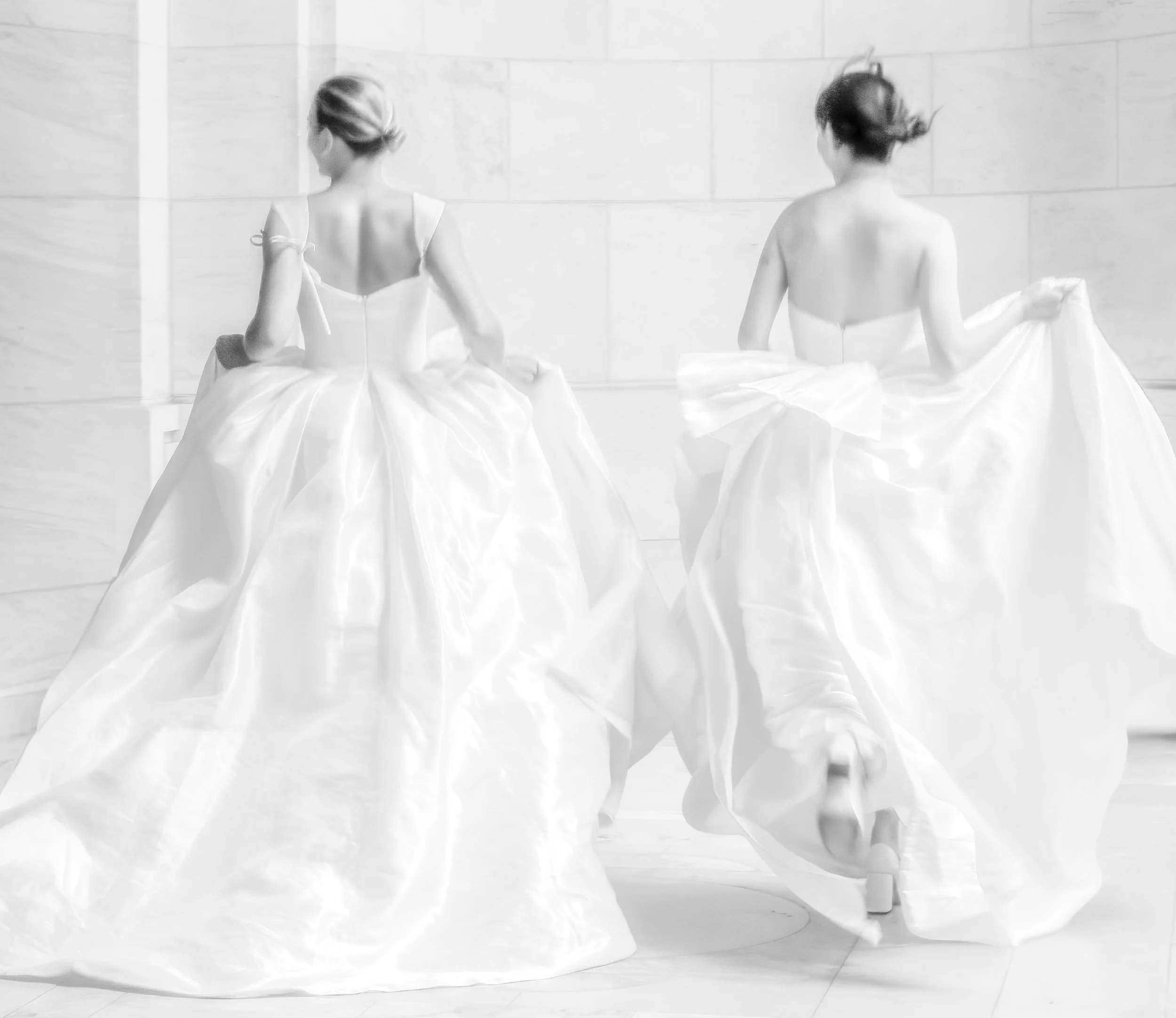 Two women in white wedding gowns posing from behind against a light stone wall