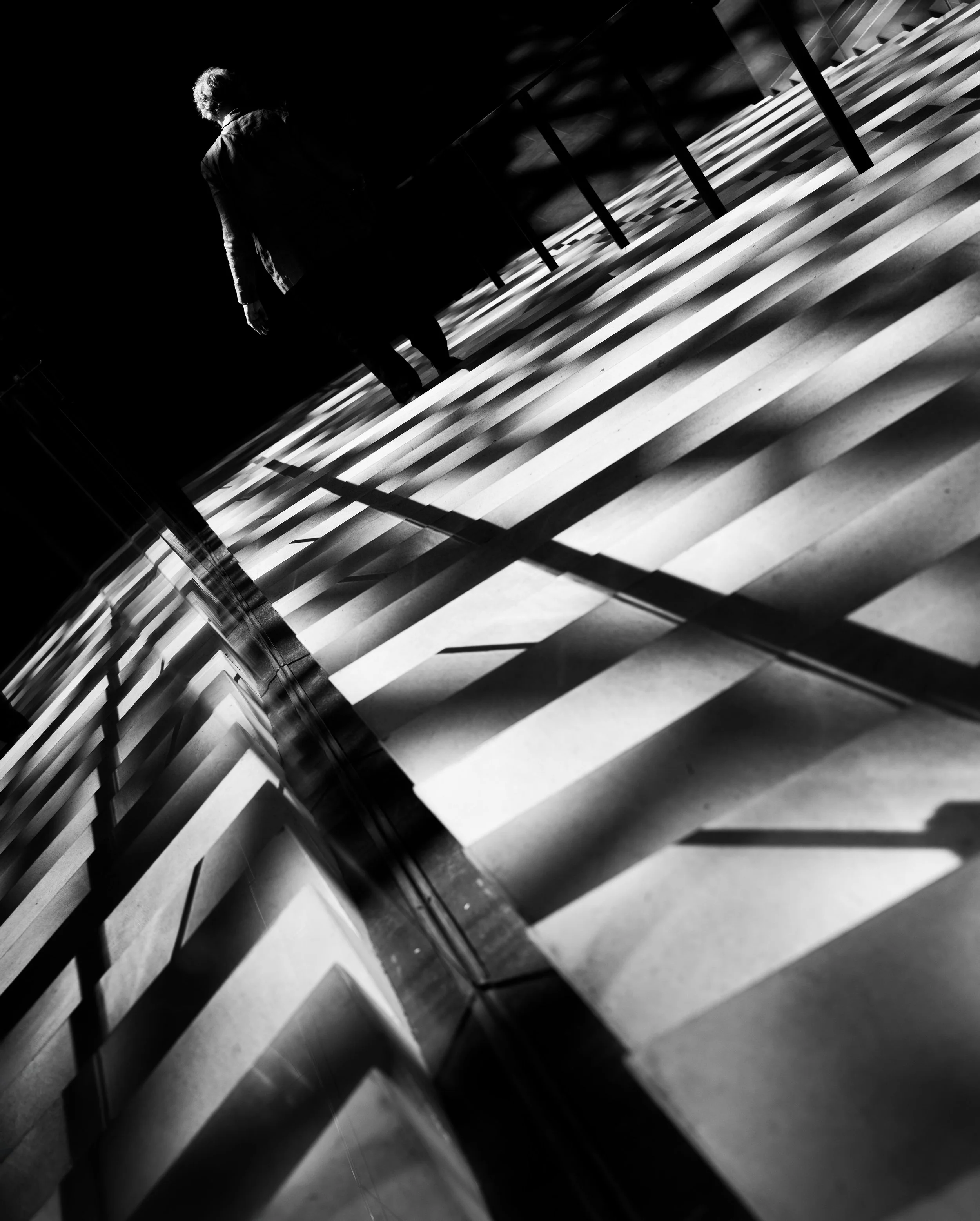 Black-and-white photo of a person walking beside a modern staircase with dramatic geometric shadows