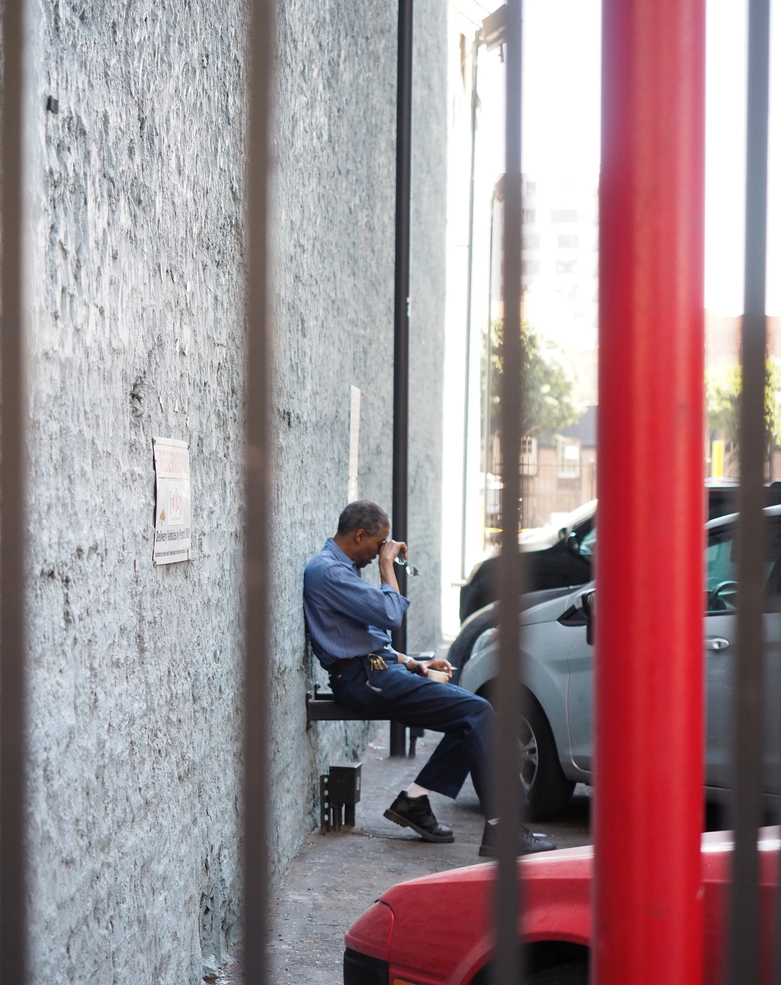 Man seated on a narrow urban sidewalk beside a stone wall and parked cars