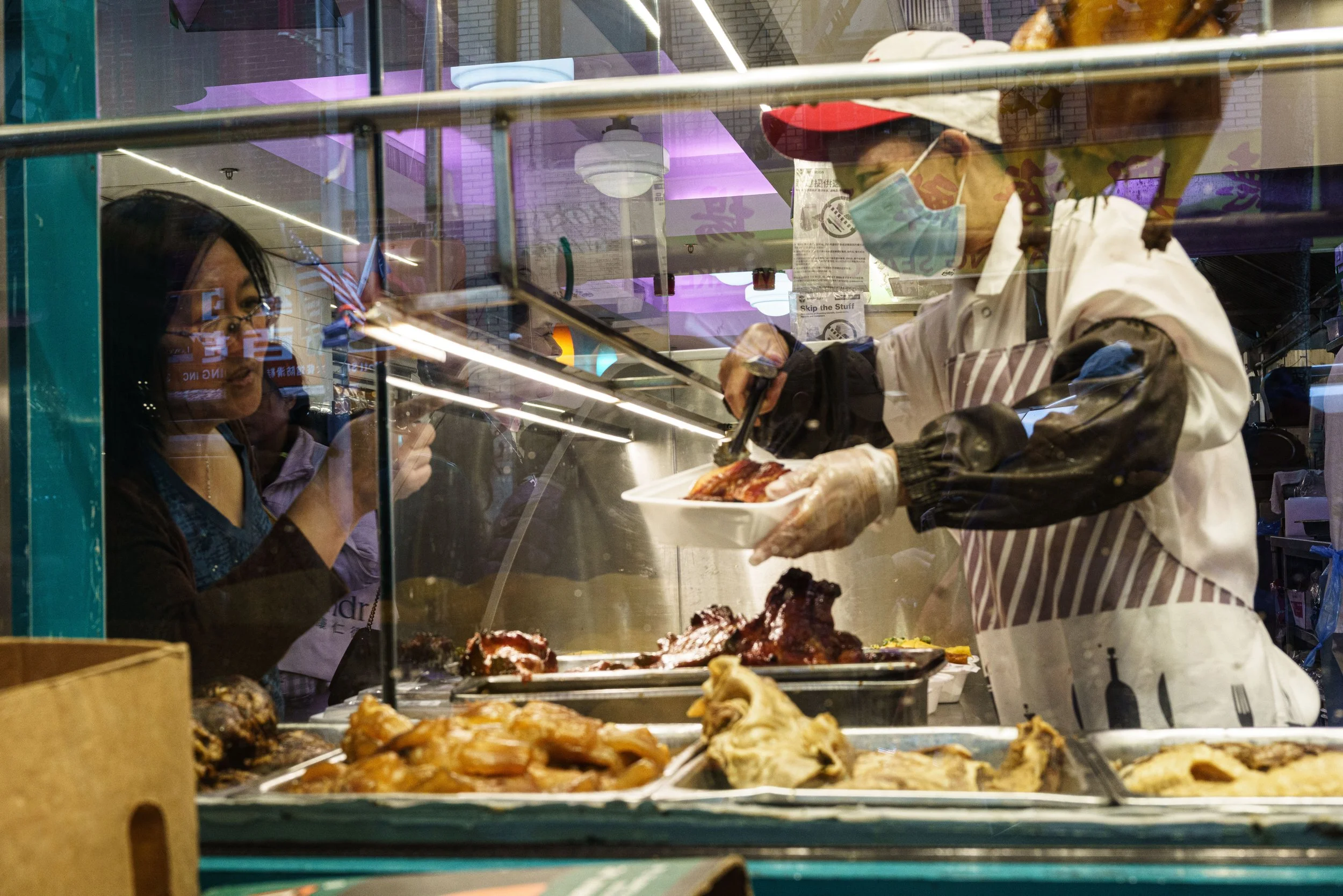 Masked vendor serving roasted meats from a Chinatown market food stall behind glass.