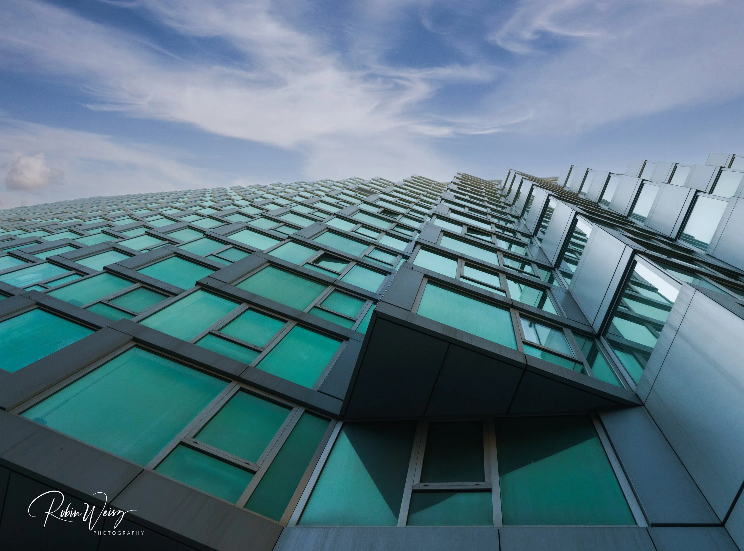 Low-angle view of a modern high-rise building with turquoise glass windows against a cloudy blue sky