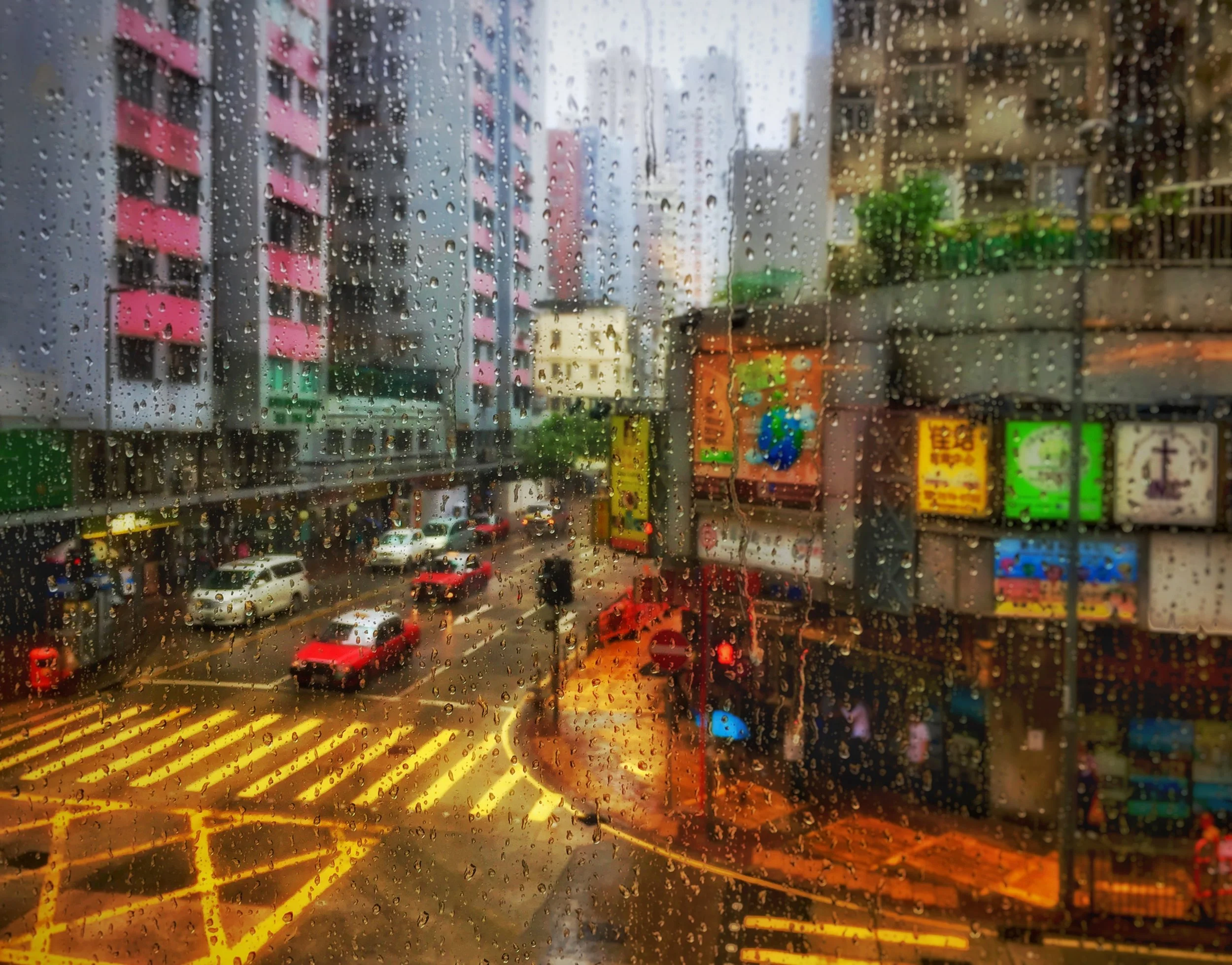 Rainy city street viewed through a window covered in raindrops, with traffic, colorful buildings, storefronts, and glowing headlights.