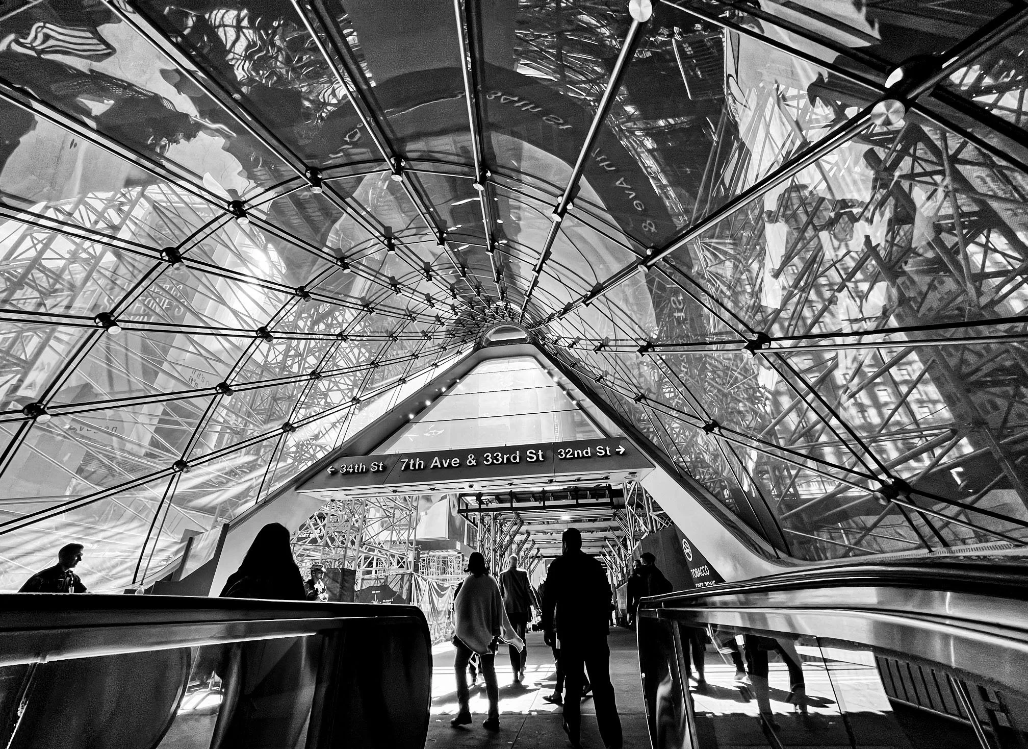 People riding escalators beneath a glass canopy at New York City’s 34th Street–Hudson Yards subway station.