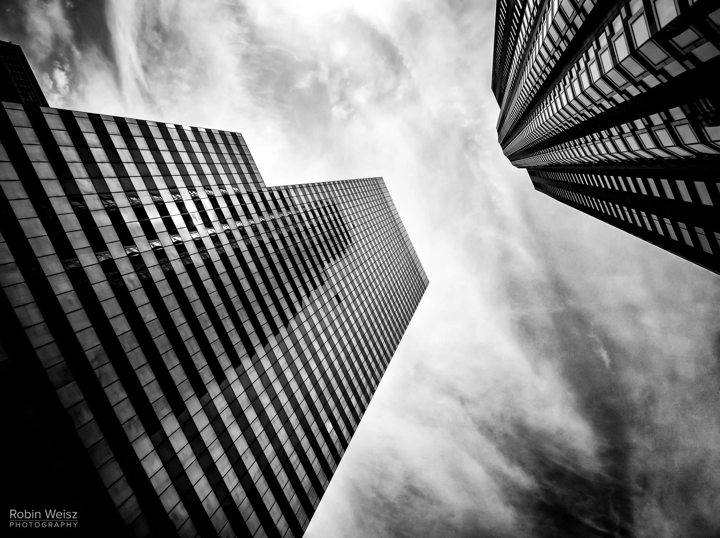 Black-and-white upward view of modern skyscrapers with geometric glass facades framing a cloudy sky