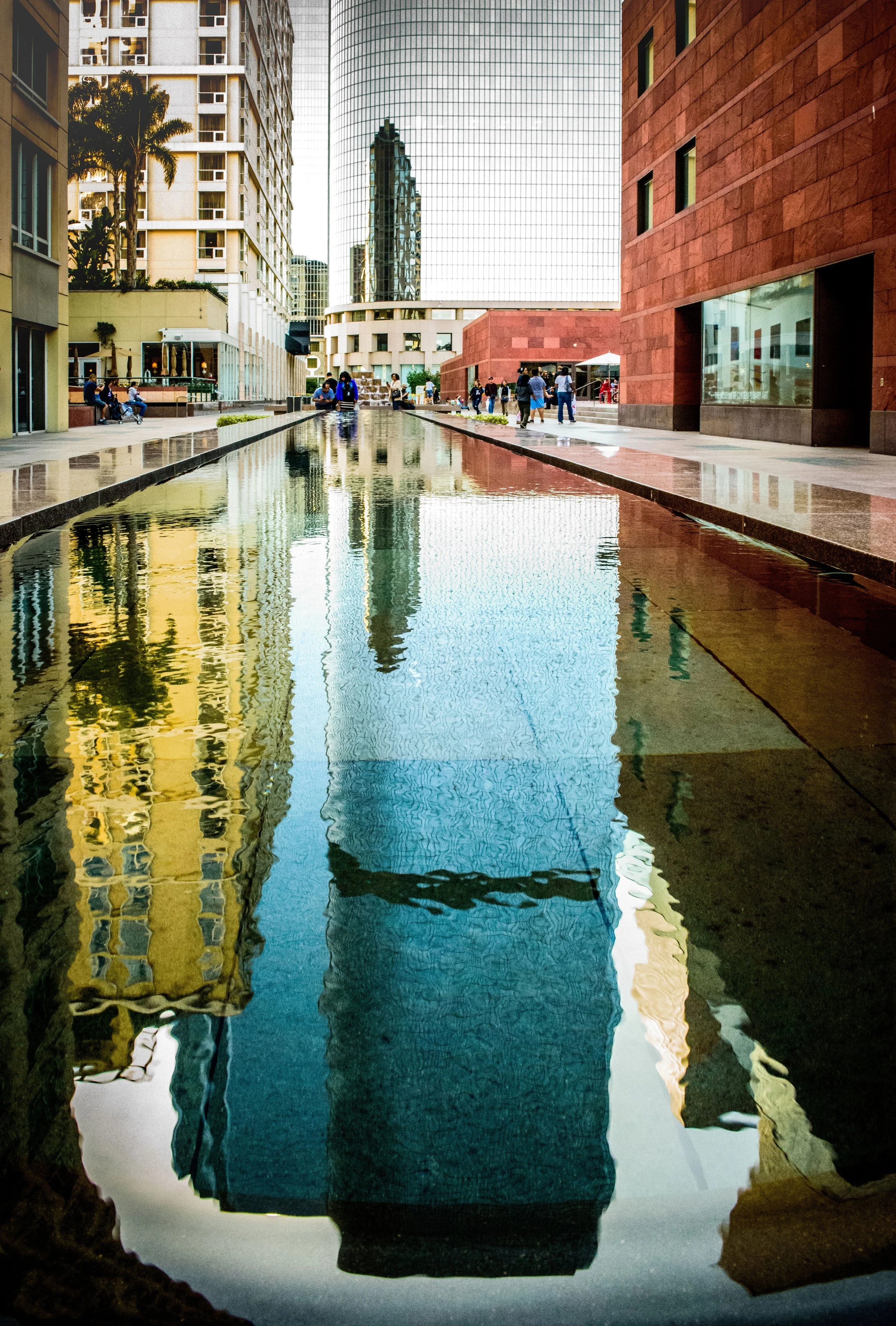 Urban shopping plaza with a reflecting pool, modern high-rise buildings, palm trees, and pedestrians