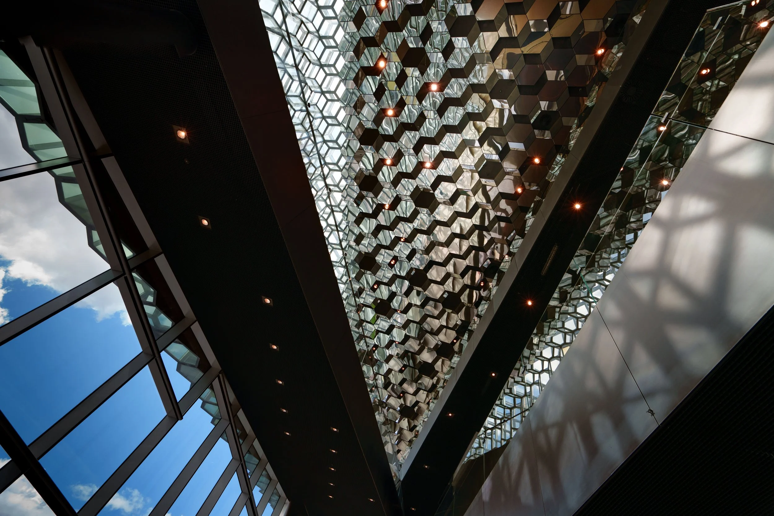 Upward view of a modern glass atrium with a honeycomb-patterned ceiling, dark beams, mirrored panels, and warm lights