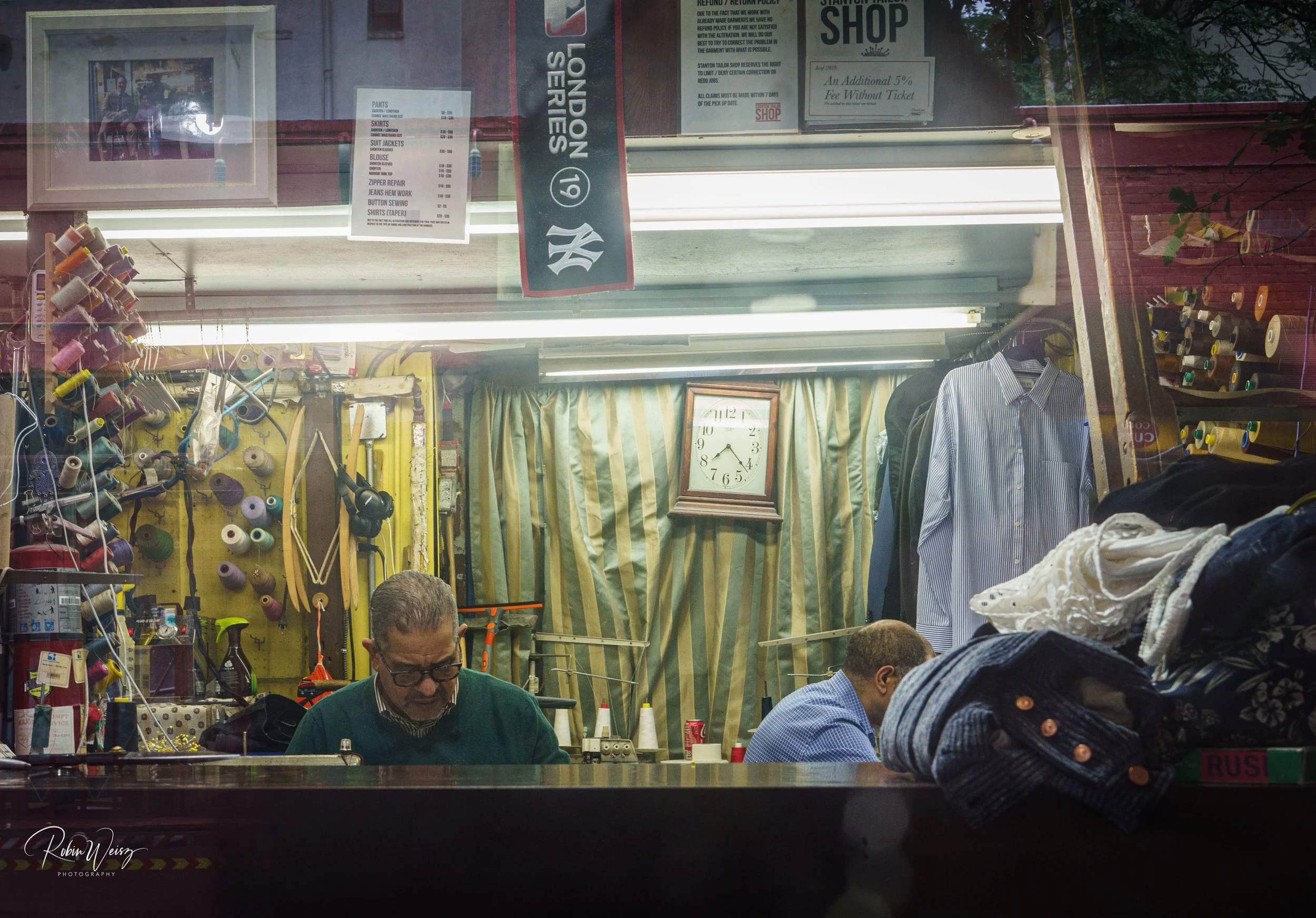 Tailor shop interior with sewing machines, colorful thread spools, clothing, alteration signs, and tailors working behind the counter