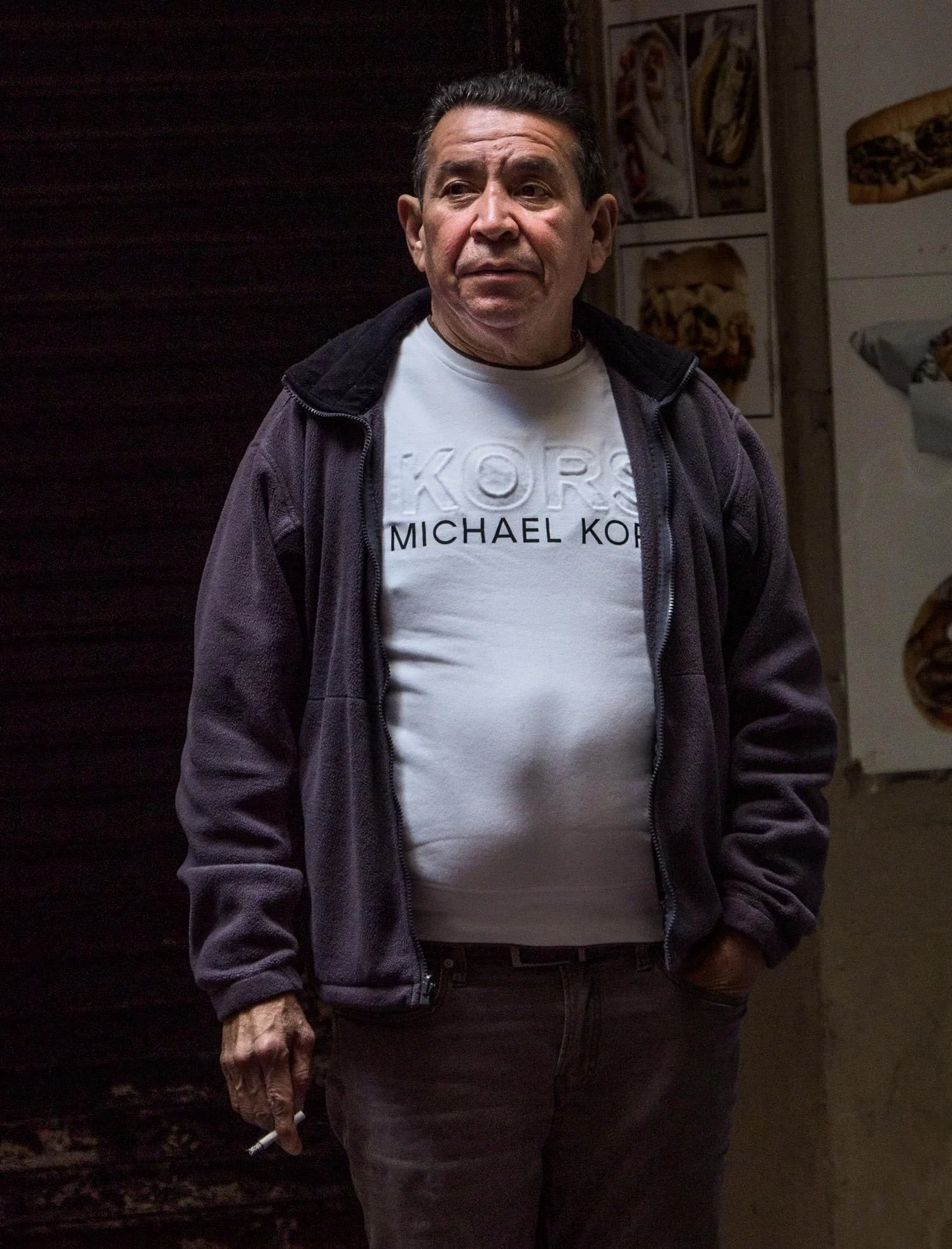 Man wearing a Michael Kors T-shirt and dark jacket, standing indoors with a cigarette.