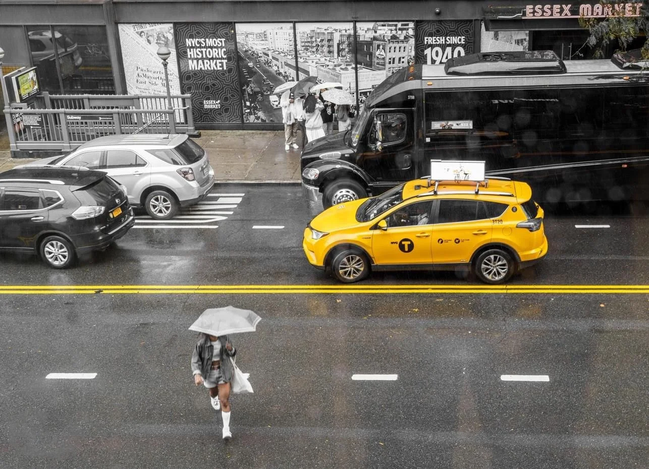 Rainy New York City street outside Essex Market, with a yellow taxi, city bus, cars, pedestrians holding umbrellas, and subway entrance.