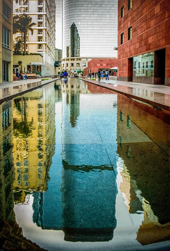 Urban plaza with a reflecting pool, palm trees, pedestrians, and modern high-rise buildings