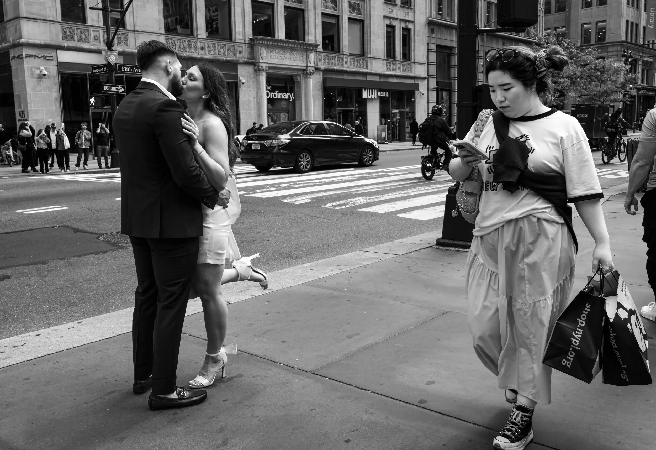 Black-and-white street scene in New York City with a couple kissing at a crosswalk and a woman checking her phone near Fifth Avenue and East 41st Street.