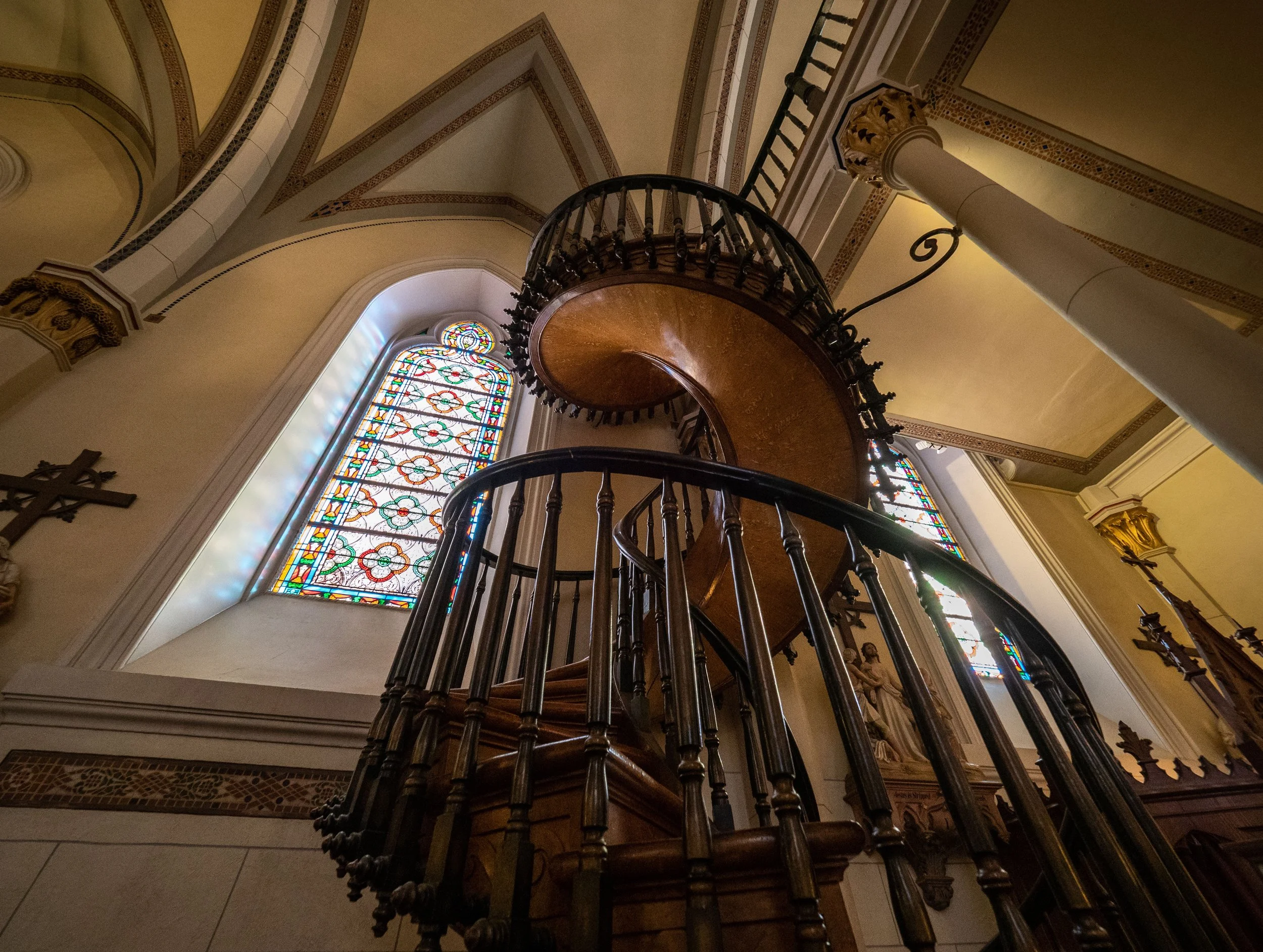 Ornate wooden spiral staircase inside a church with stained-glass windows and decorative vaulted architecture
