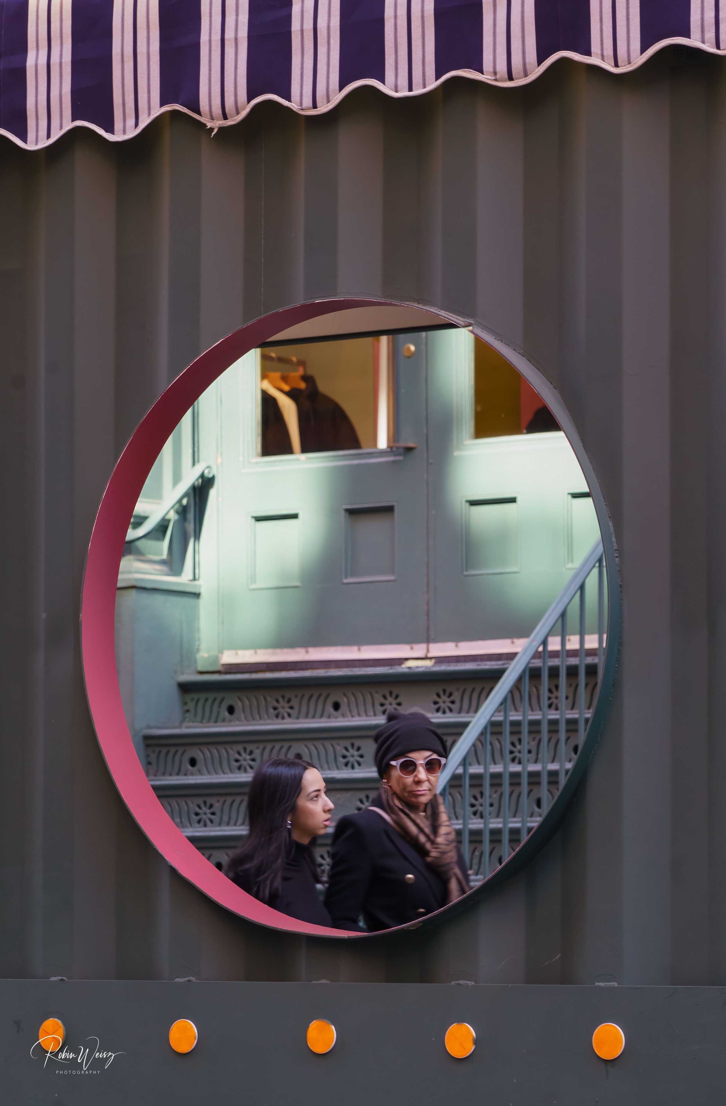 Two women reflected in a large circular storefront mirror beneath a striped awning.