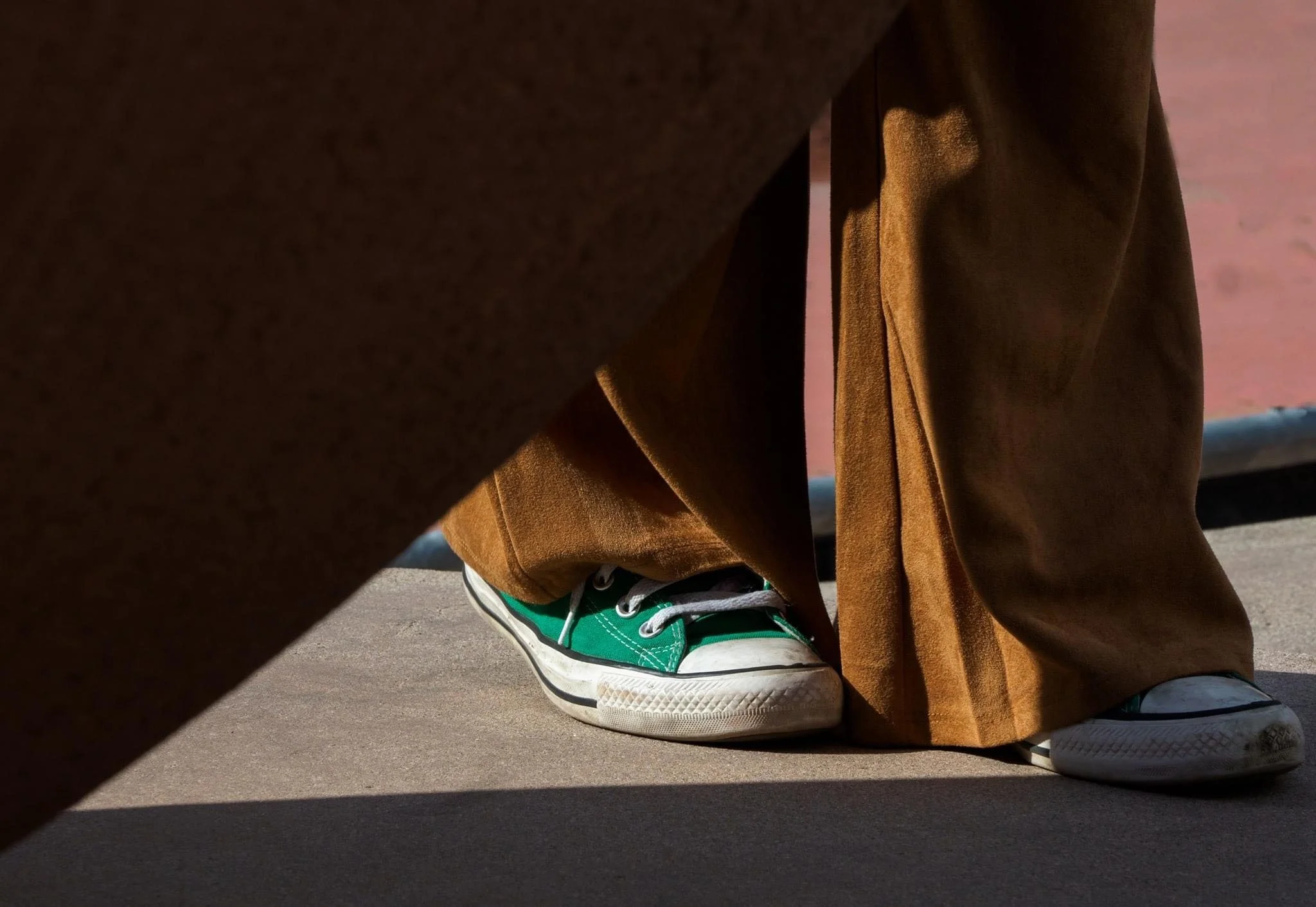 Close-up of brown wide-leg pants and green canvas sneakers on a sunlit pavement.