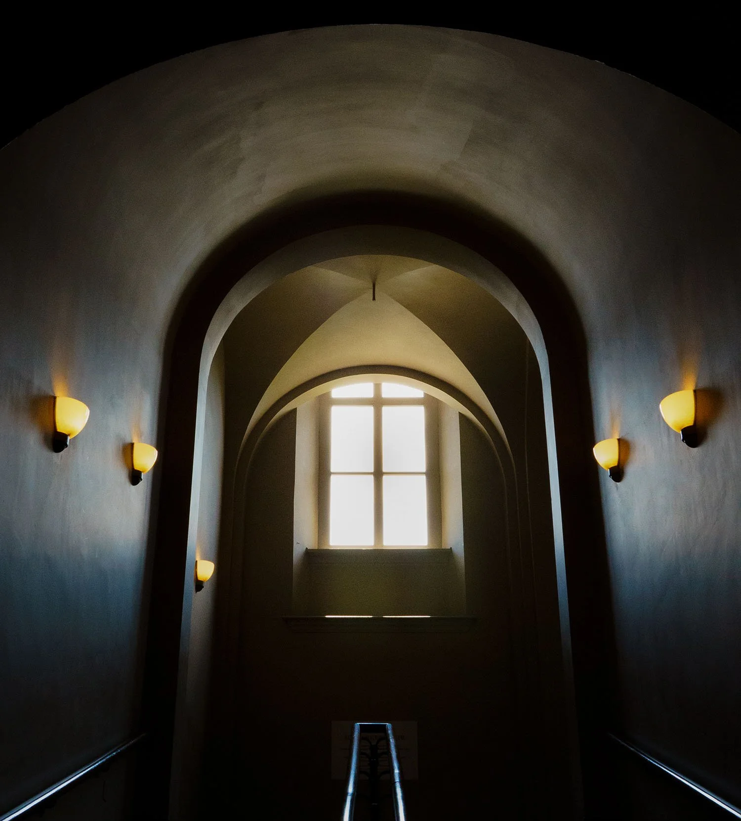 Dimly lit arched interior hallway with glowing wall sconces and a large cross-shaped window.