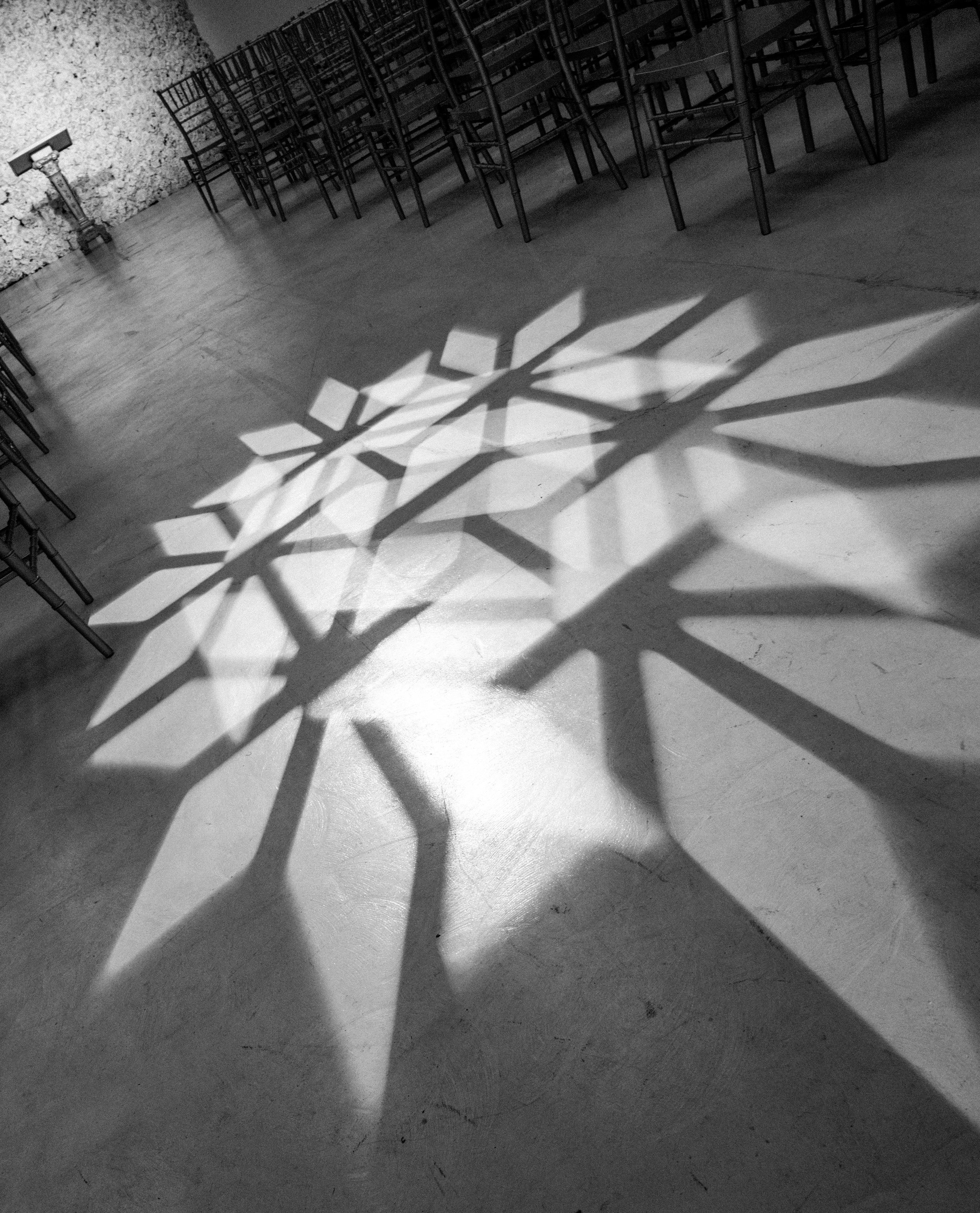 Black-and-white interior with rows of wooden chairs casting long geometric shadows across a concrete floor.