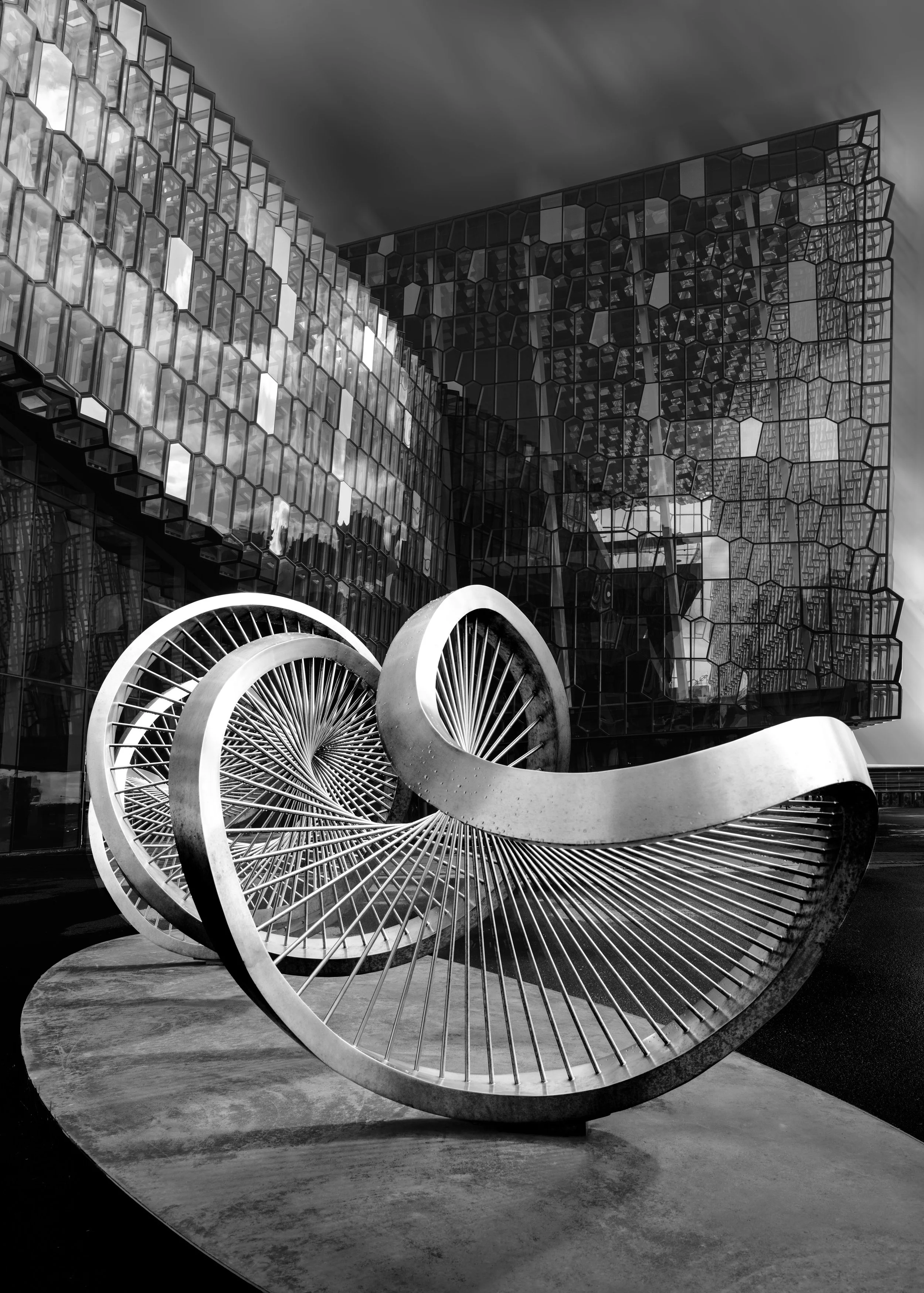Black-and-white photo of an abstract metal sculpture with curved rings and radiating spokes in front of a geometric glass building.