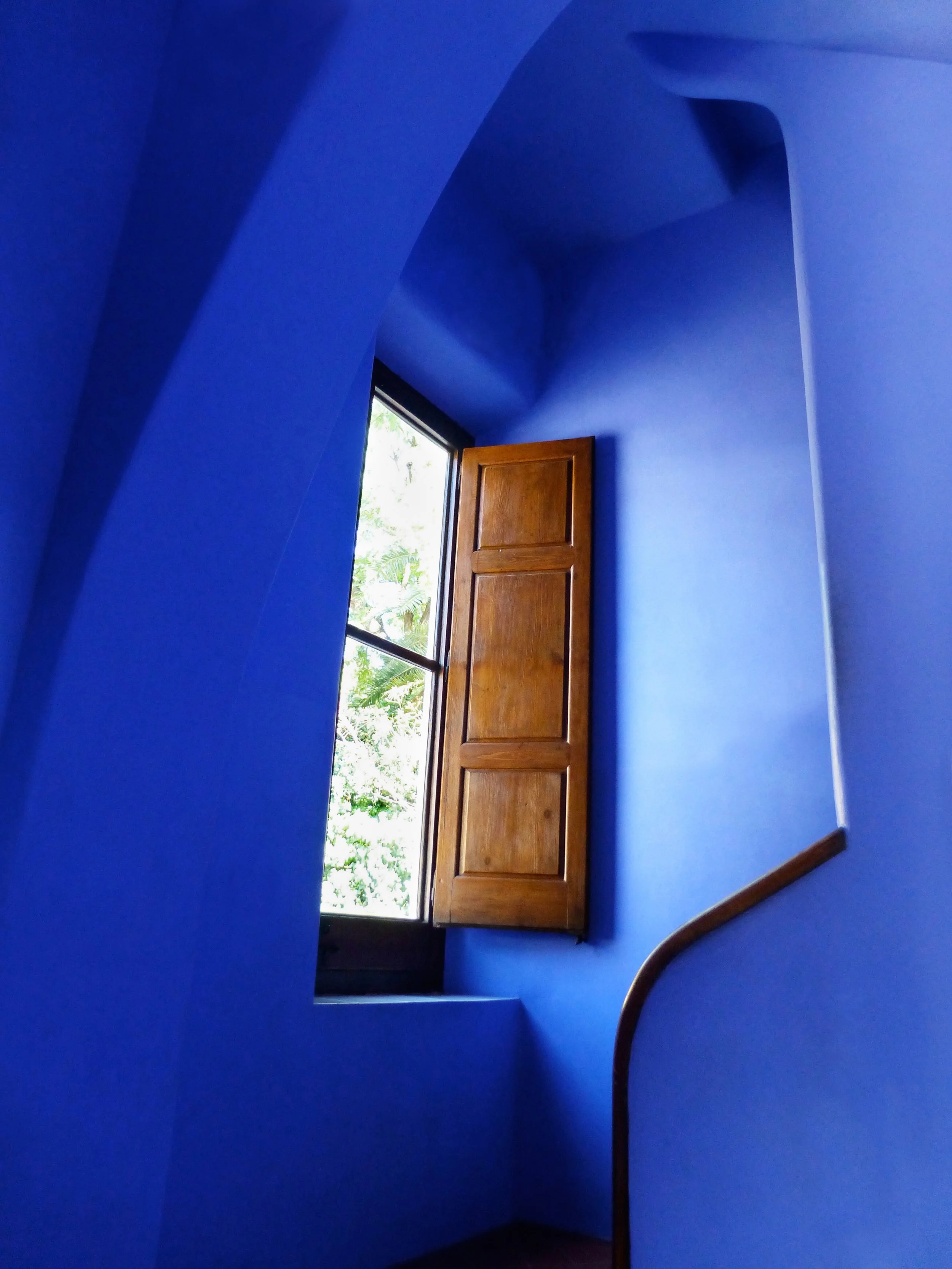 Bright blue room with an open wooden window, tropical greenery outside, and a curved wooden railing.