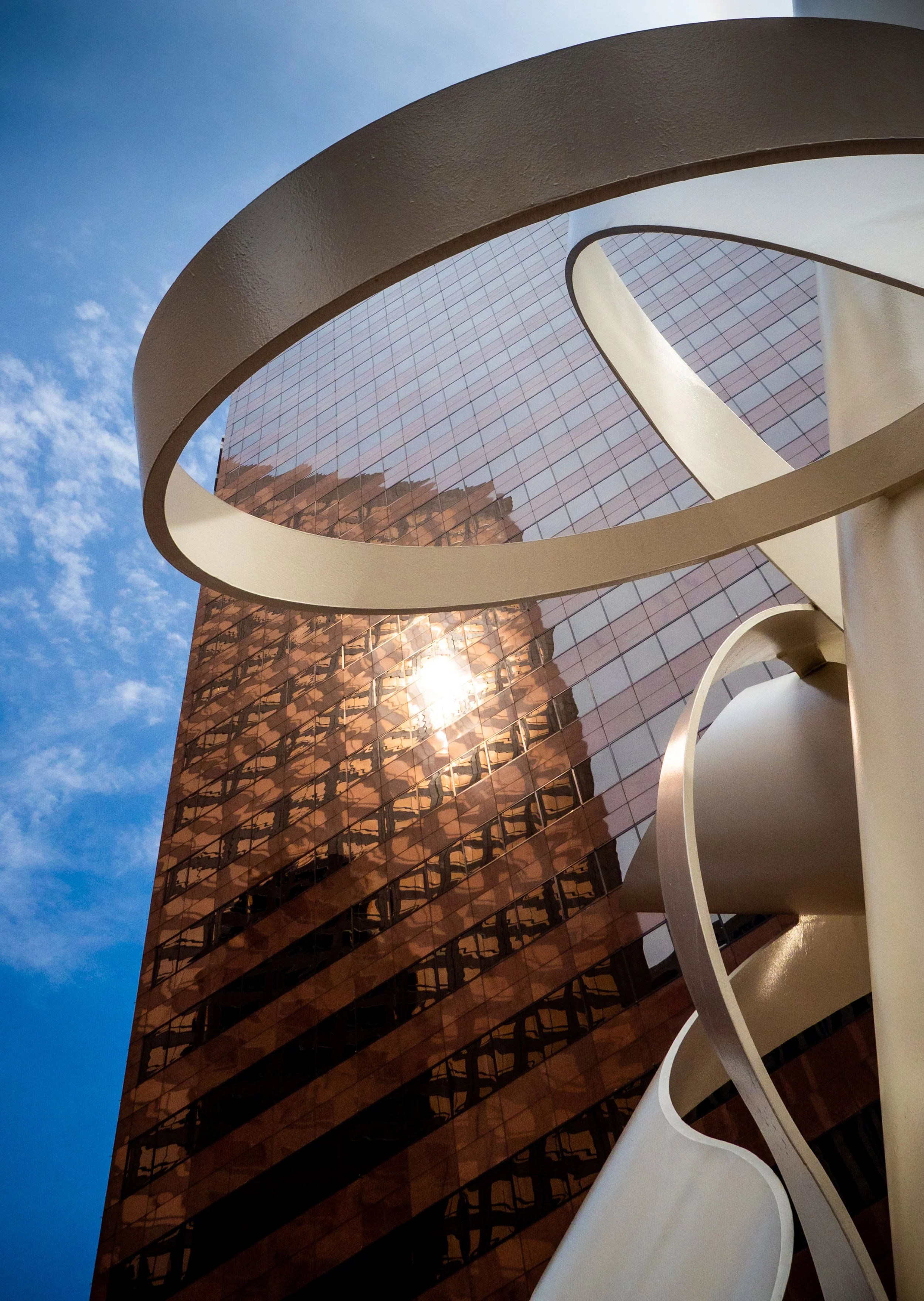 Upward view of a modern glass skyscraper framed by sweeping white architectural arches against a blue sky