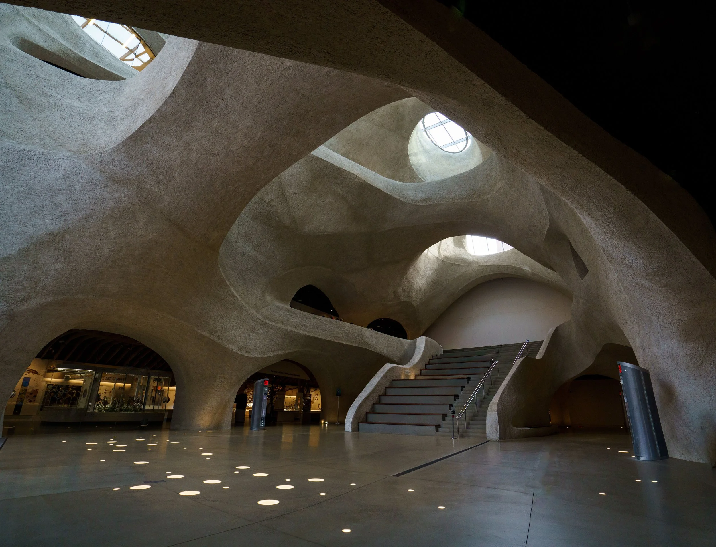 Interior of a sculptural concrete building with curved walls, skylights, archways, and a central staircase.