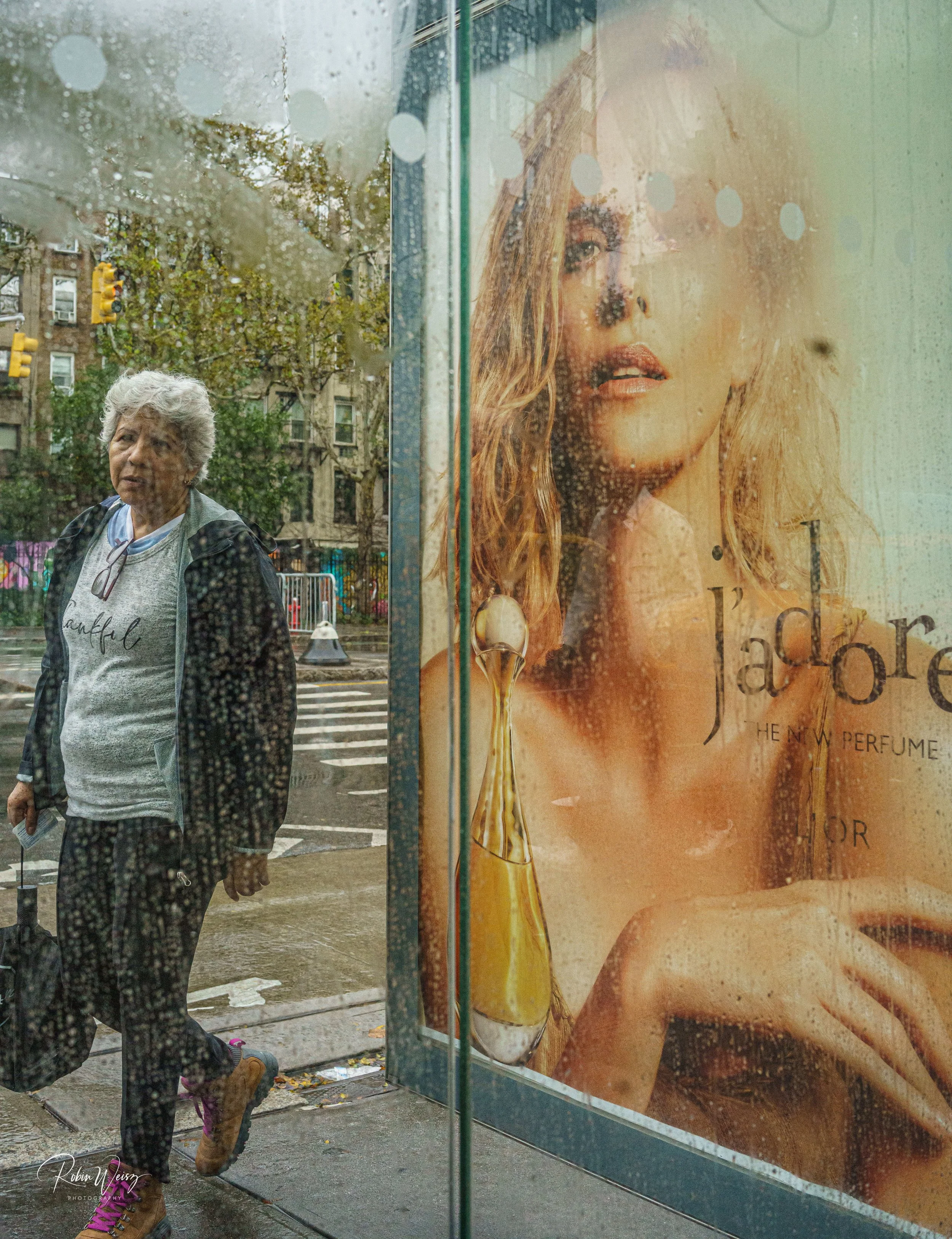 Older woman walking past a rain-speckled shop window displaying a large perfume advertisement on a city street.