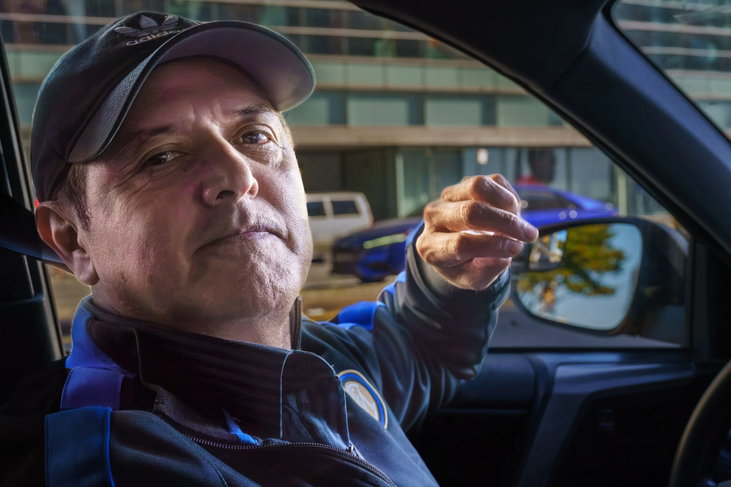 Man wearing a cap and blue jacket sitting inside a car, gesturing toward the camera