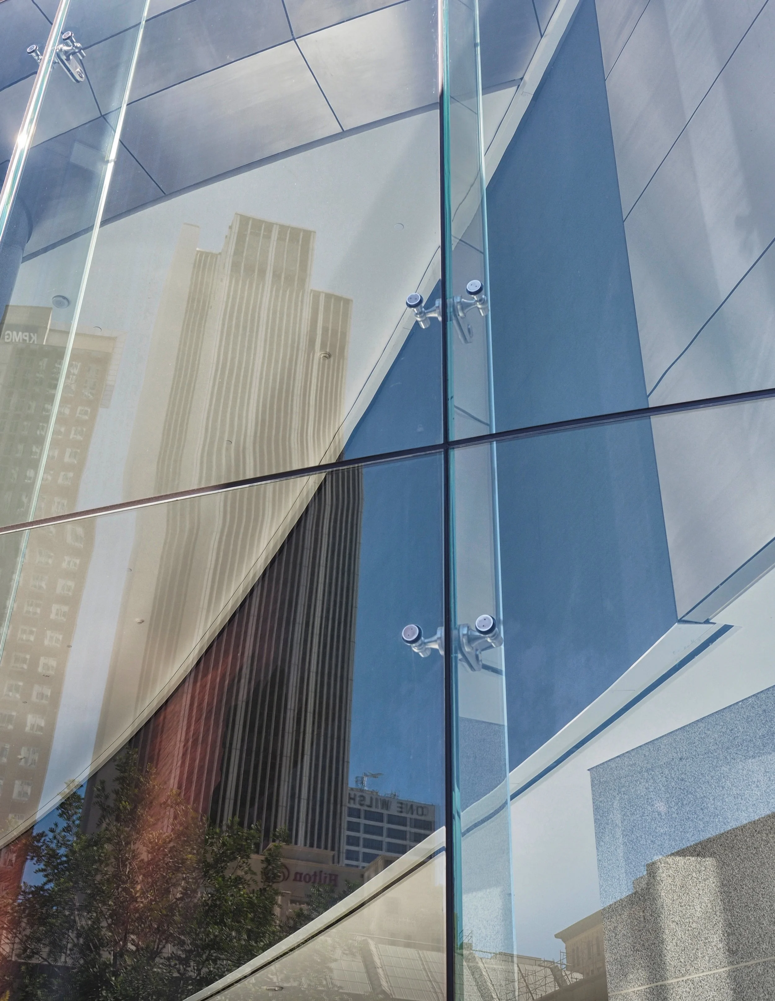 Close-up of a modern glass building facade with metal glass clamps, geometric reflections, and nearby city buildings and trees.