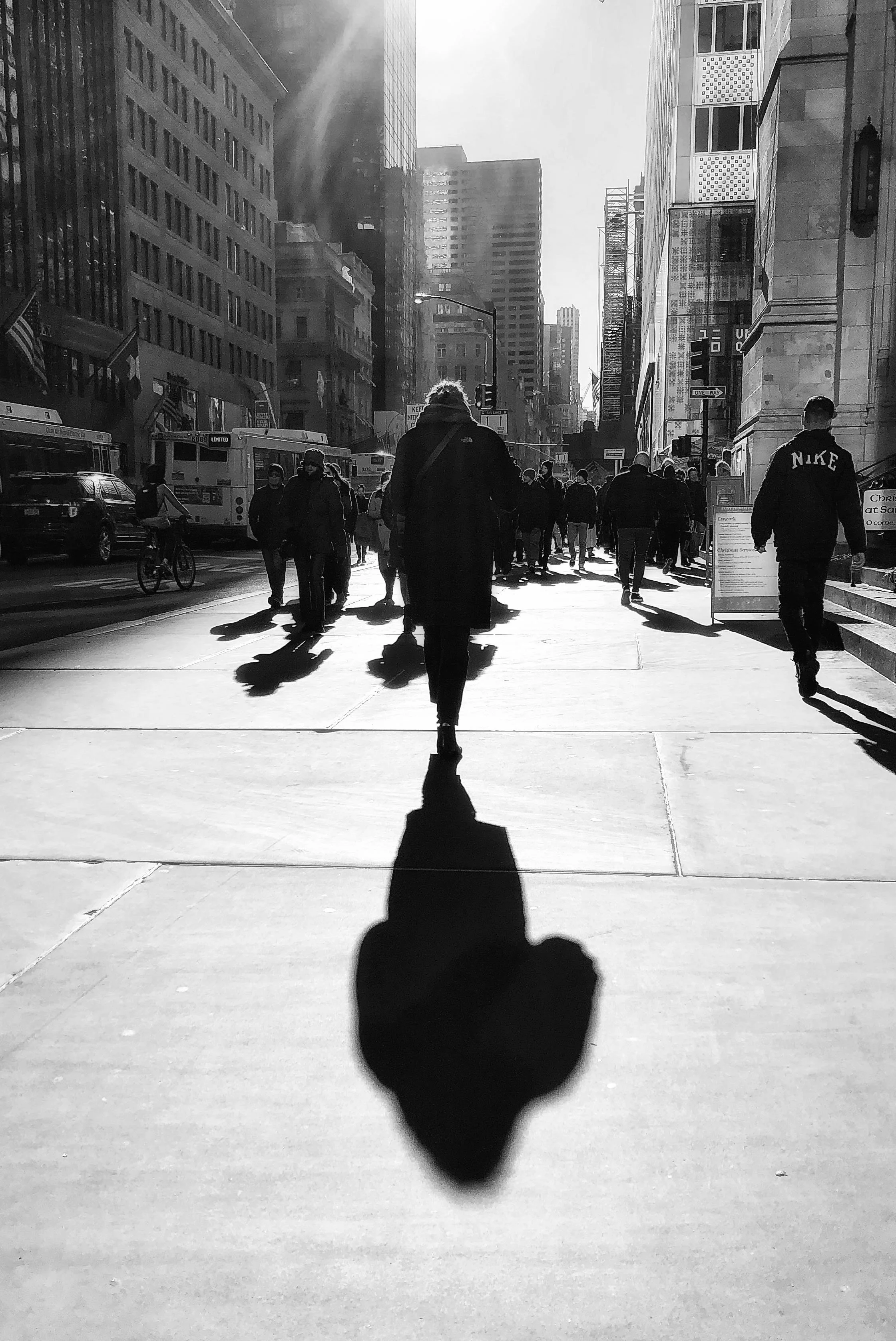 Black-and-white street scene with pedestrians, city buses, skyscrapers, and long shadows on a sunny urban sidewalk