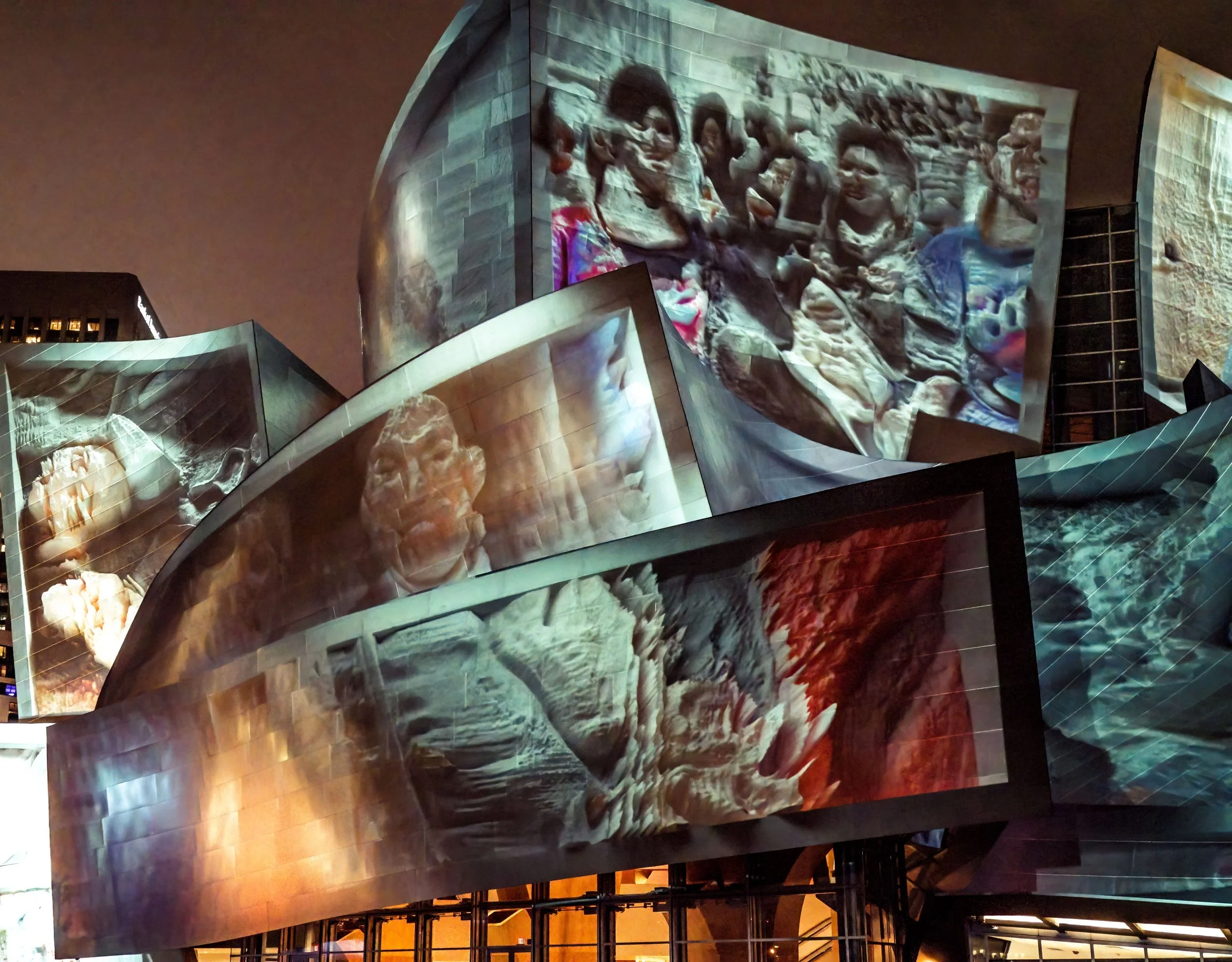 Large-scale illuminated projection of colorful human figures and abstract imagery on angular building panels at night.