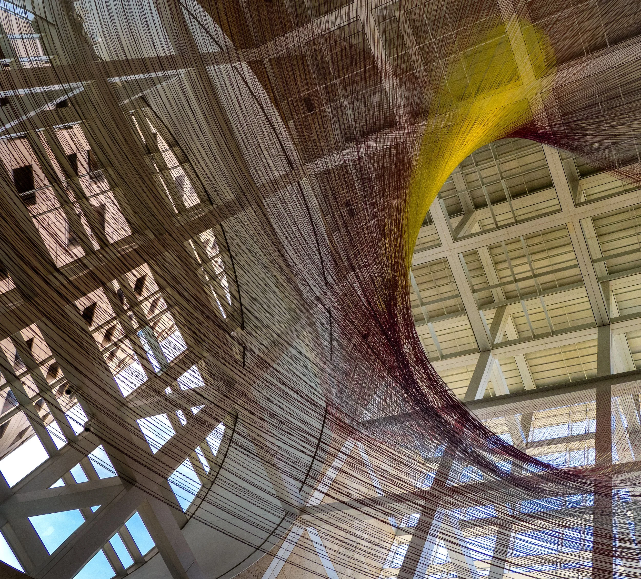 Abstract installation of dense burgundy and yellow wires suspended inside a modern glass-and-steel atrium