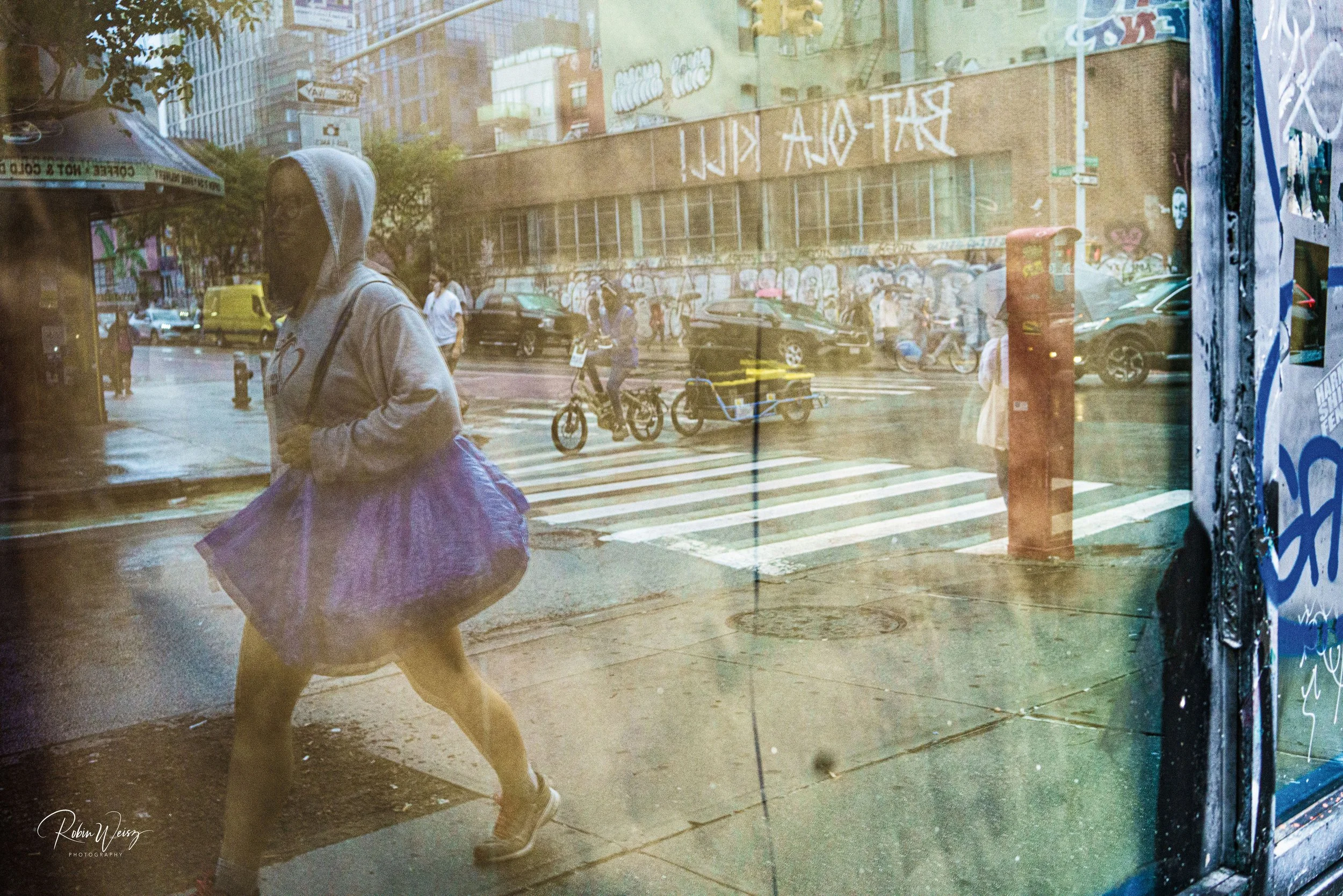 Person in a gray hoodie walking past a graffiti-covered city street, reflected in a storefront window