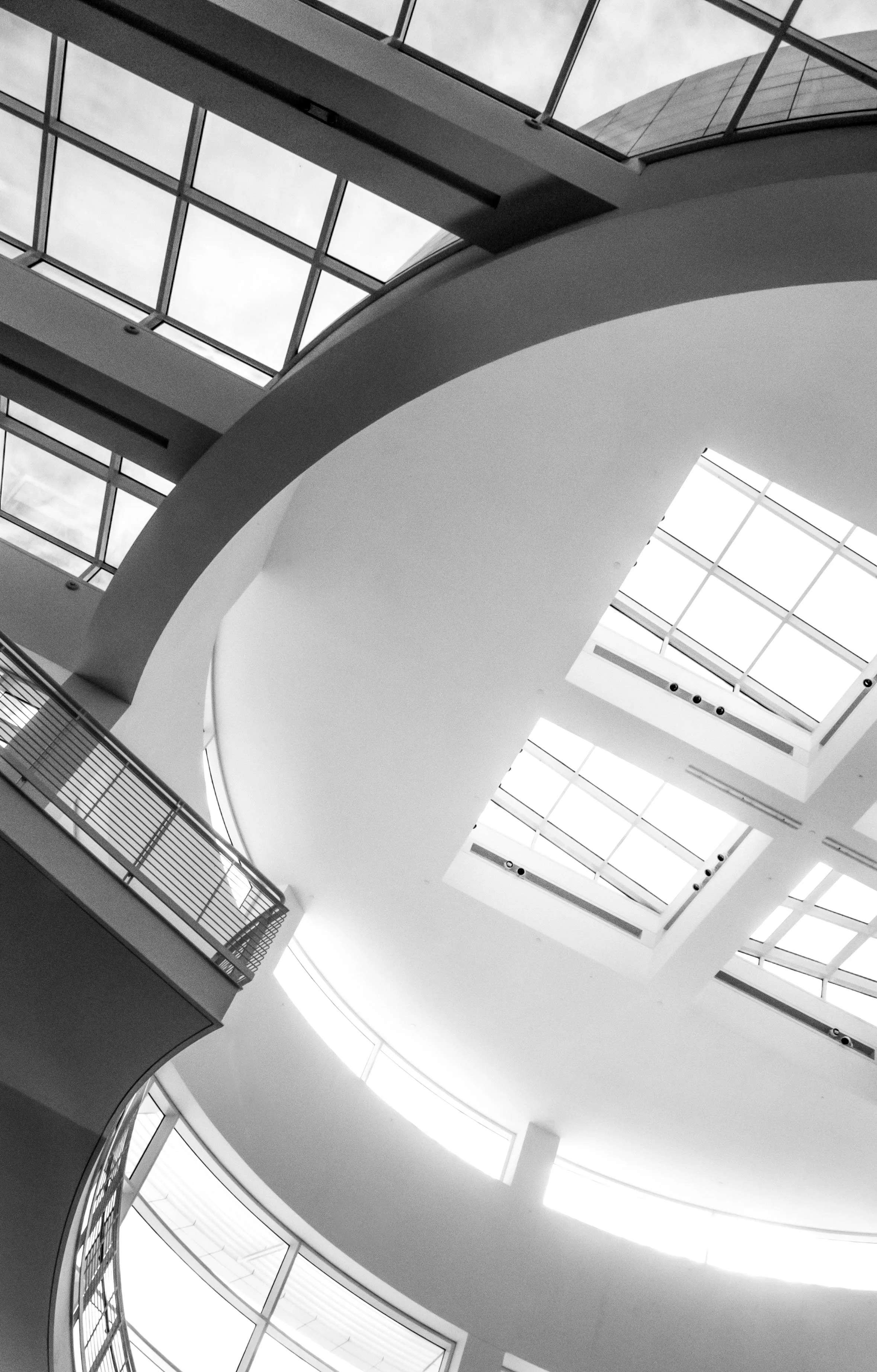 Black-and-white architectural view of a modern atrium with skylights, structural beams, white columns, and a metal balcony railing.